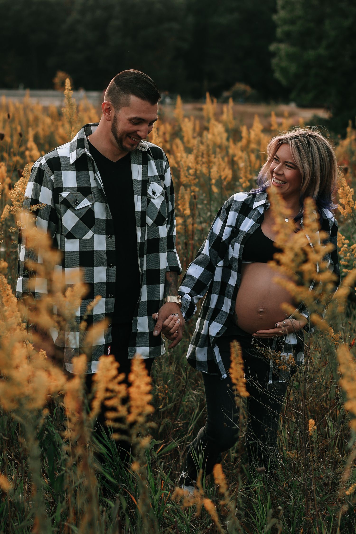 Happy couple in matching plaid shirts holding hands laughing in golden autumn wildflower field.
