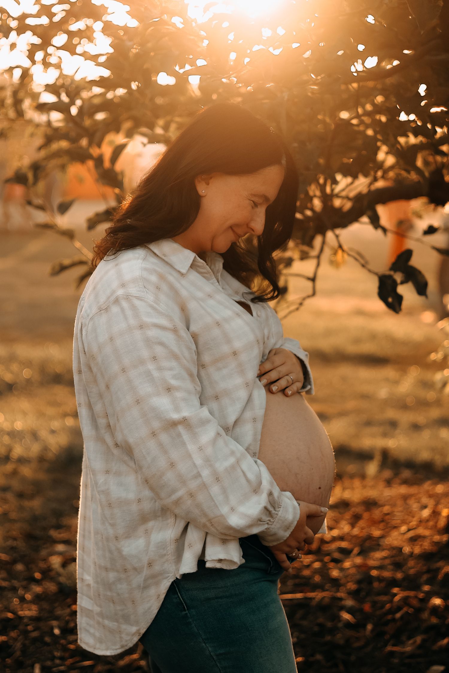 Smiling pregnant woman in plaid shirt cradling her bare baby bump outdoors in warm golden sunset light.