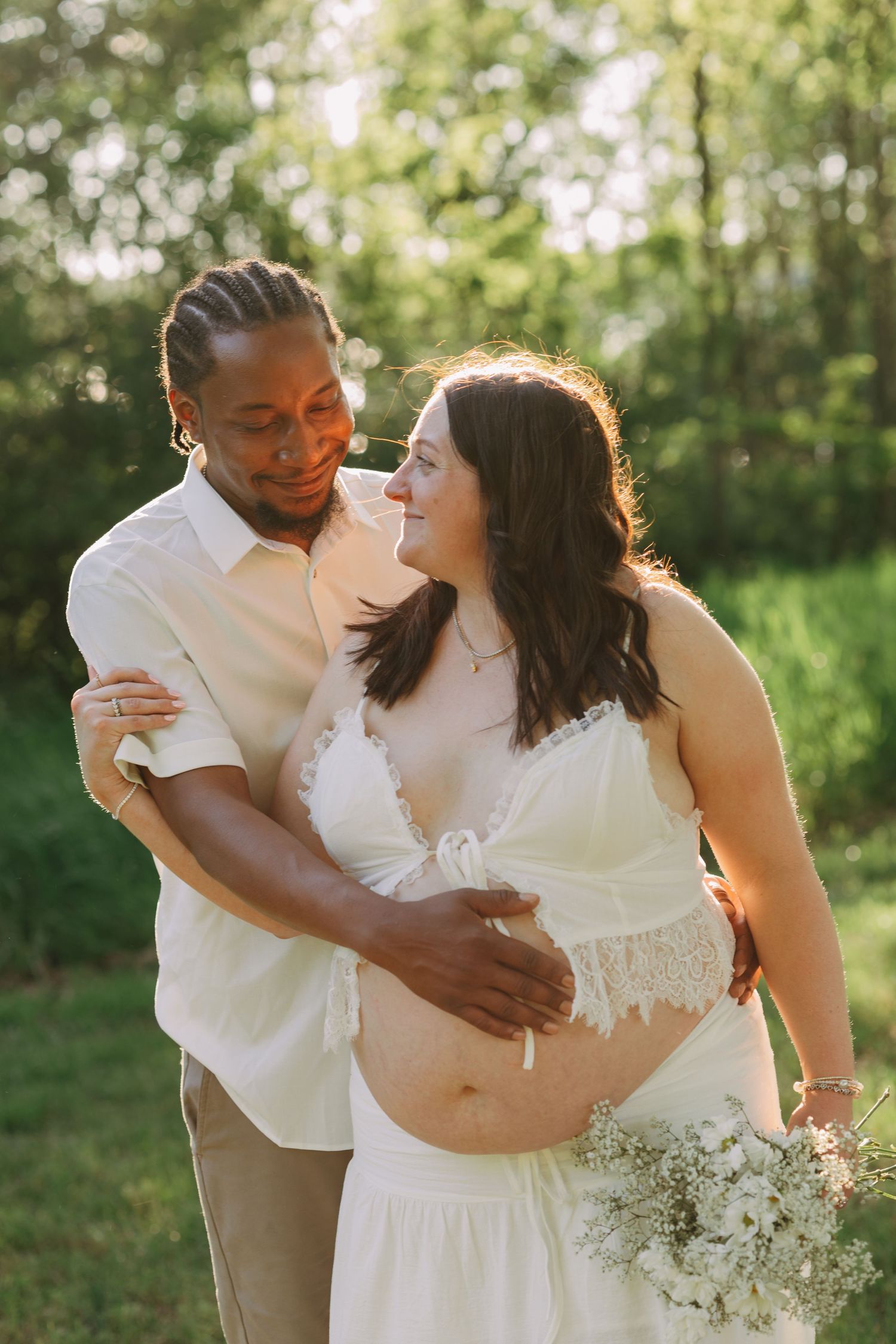 Interracial couple sharing a tender moment outdoors, pregnant woman in white lace dress holding baby's breath flowers.