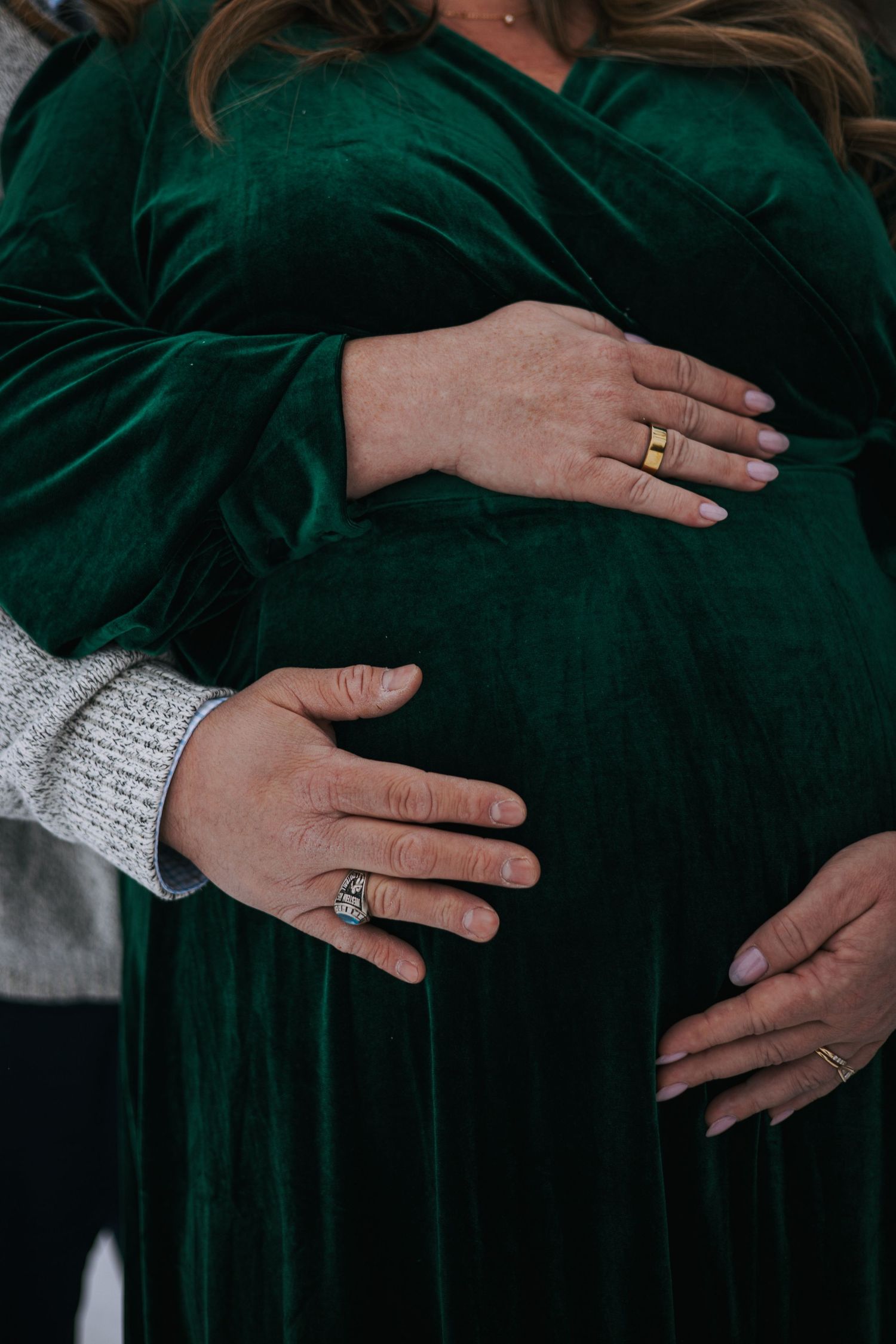 Close-up of couple's hands cradling pregnant belly in green velvet dress with wedding rings visible.