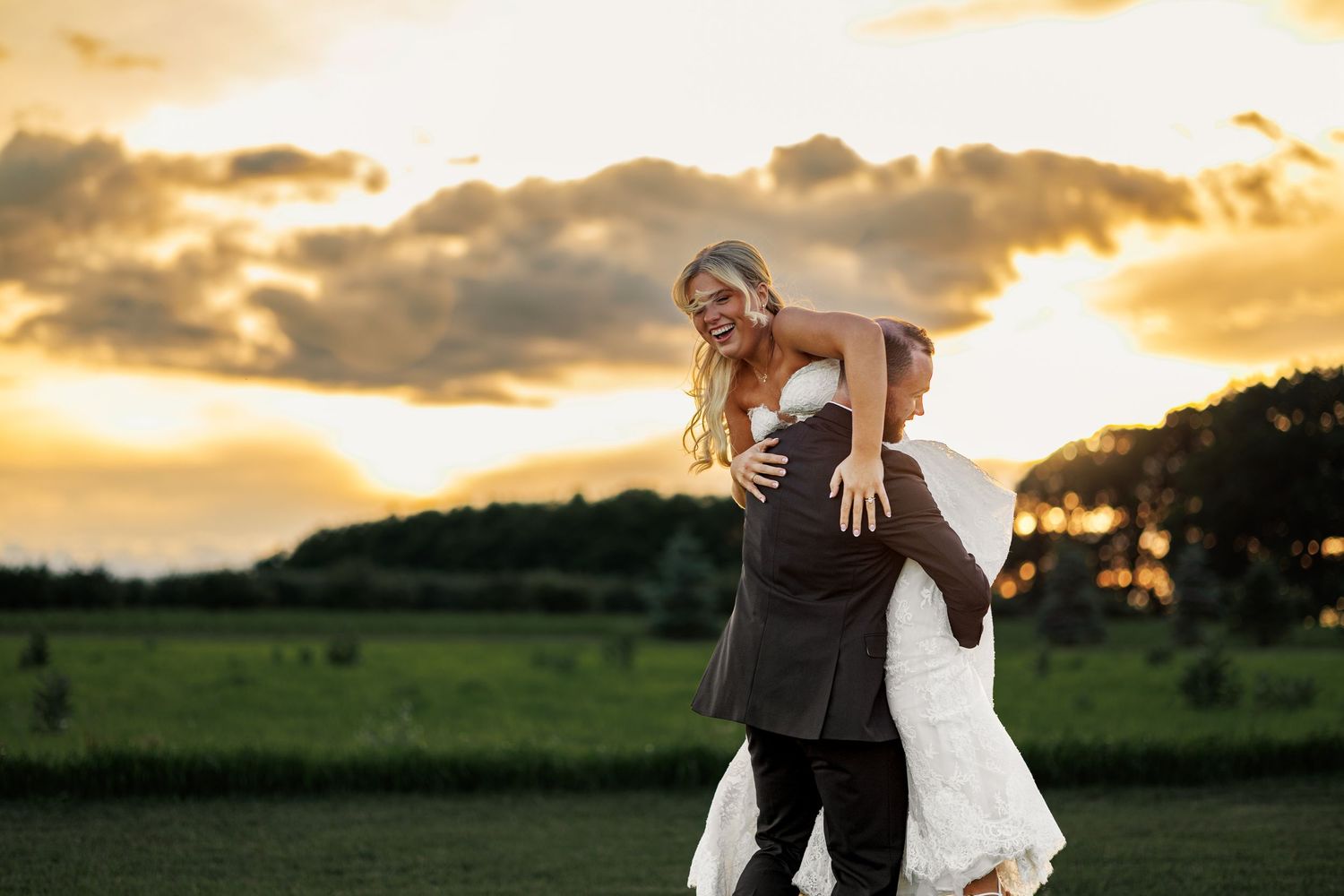 Groom lifting bride in white dress at sunset with green fields and dramatic clouds in background.