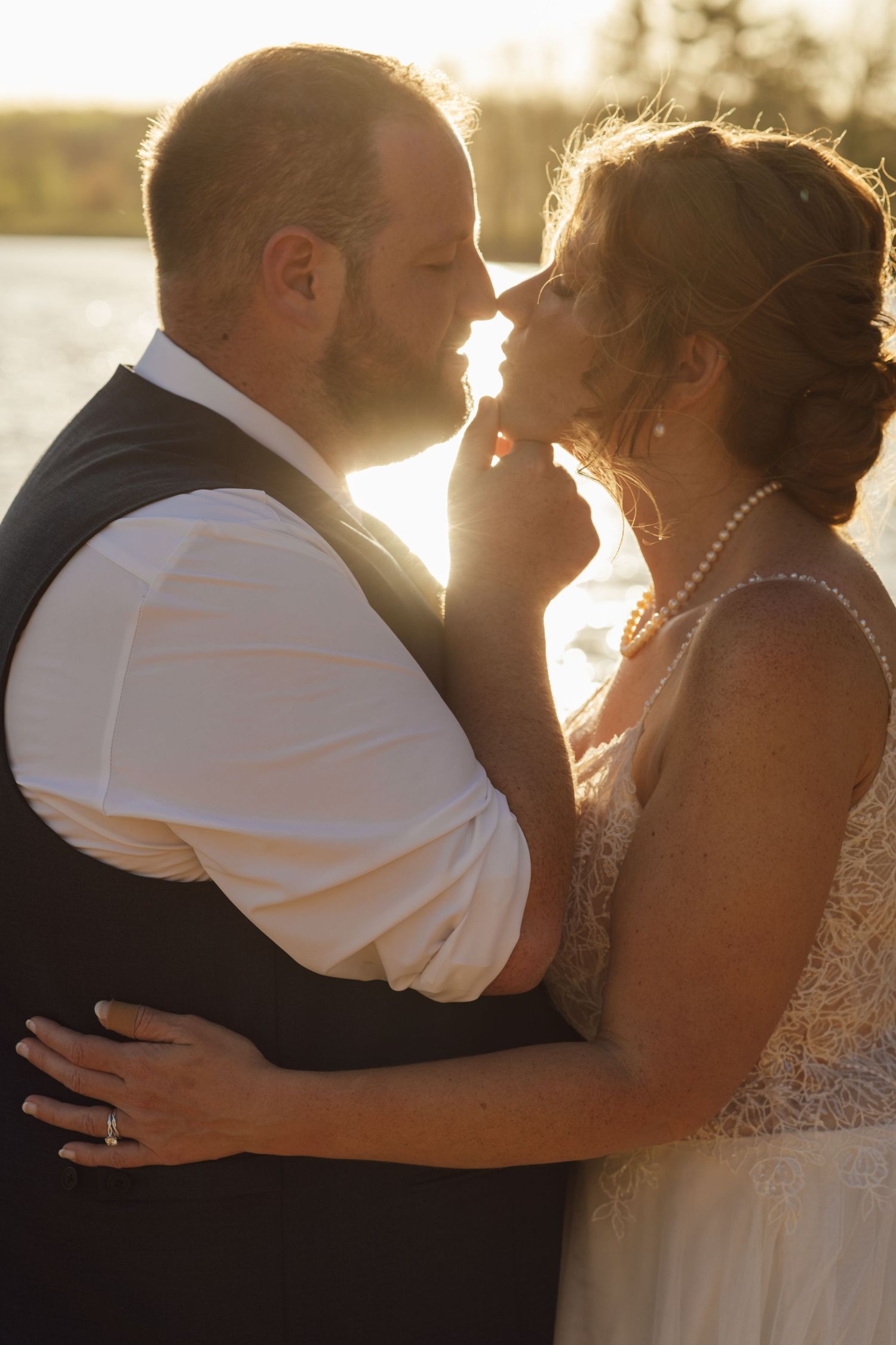 Bride and groom share a romantic moment at golden hour sunset, with bride in lace gown and pearl necklace.
