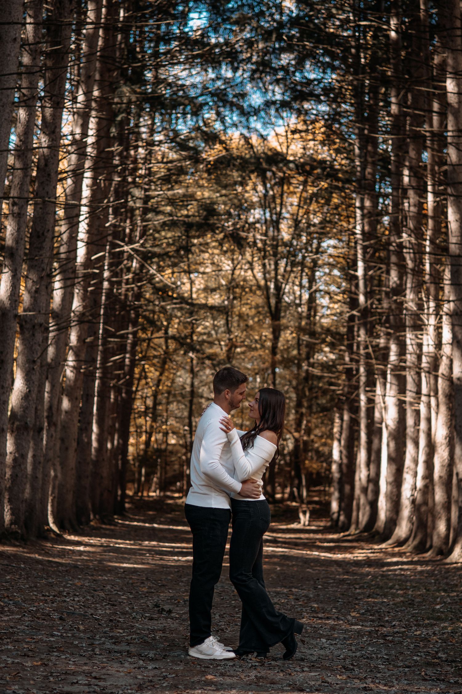 Couple embracing face-to-face on a forest path surrounded by tall trees in autumn.