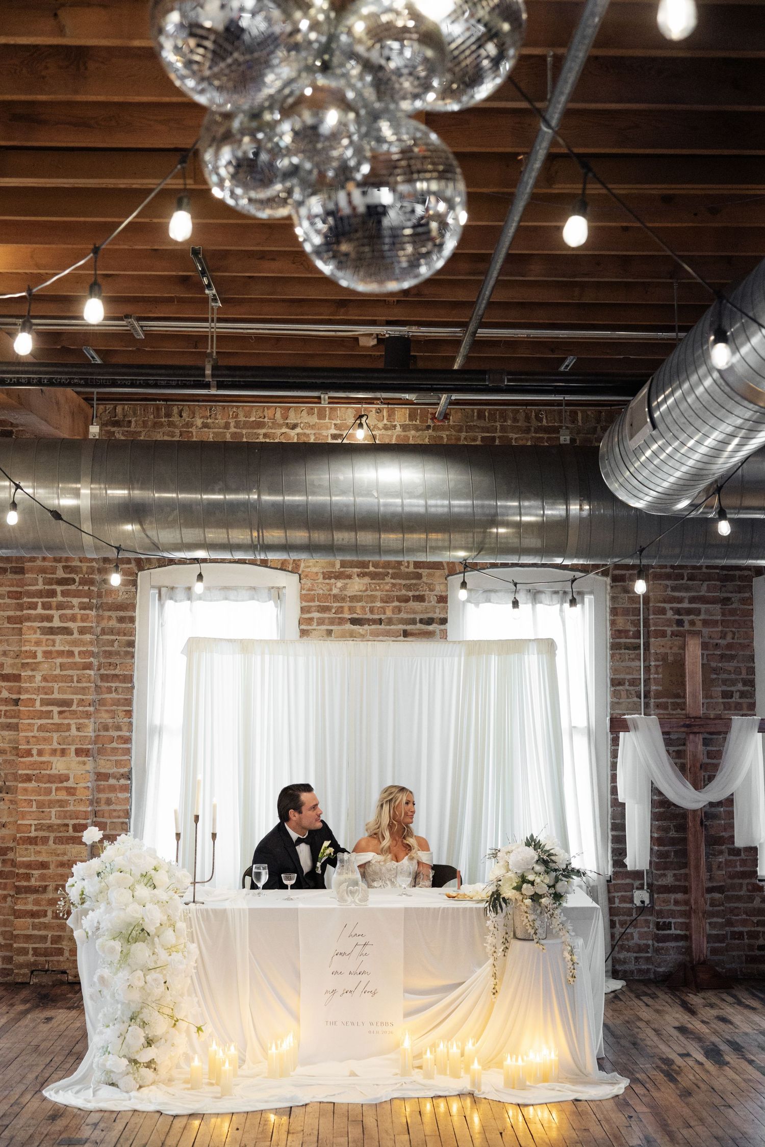 Bride and groom sit at floral sweetheart table under disco balls in a brick venue.