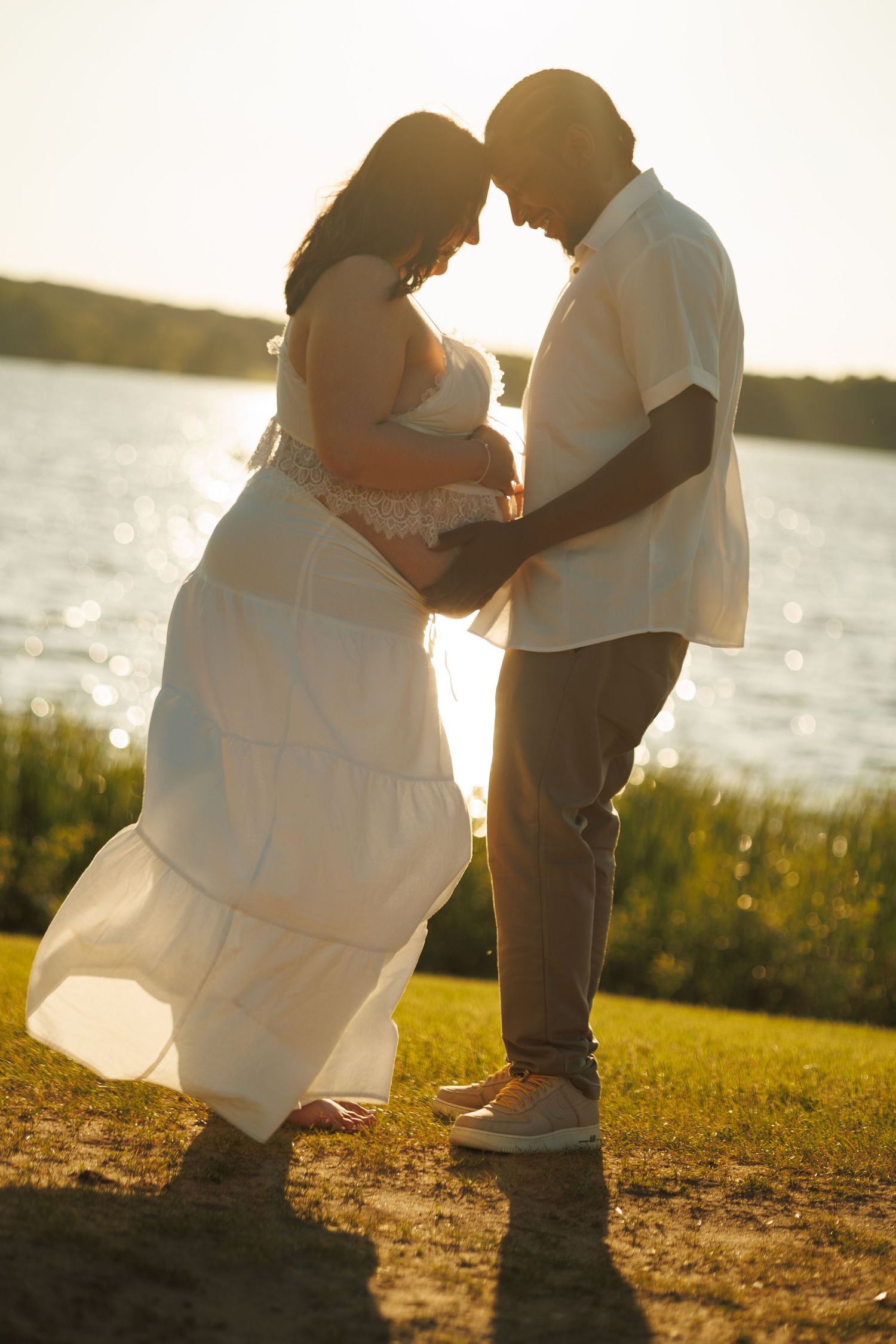 Couple sharing a tender moment at sunset by a lake, woman in white dress with hands on baby bump.