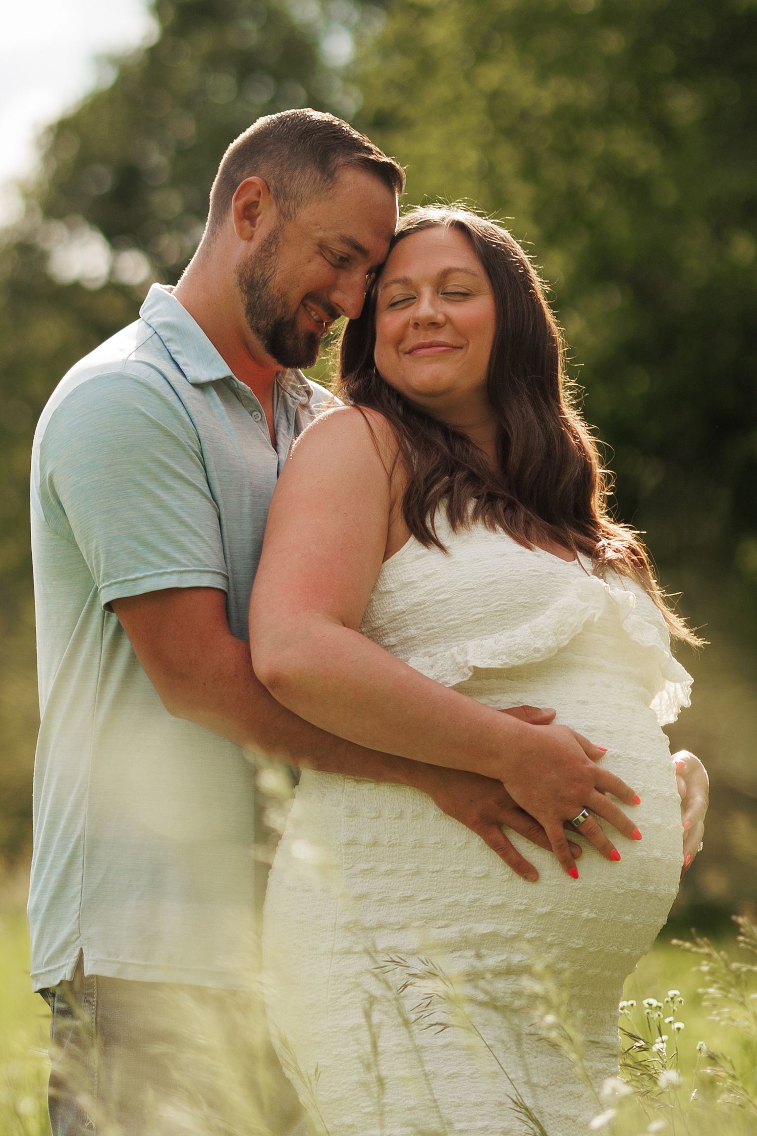 A couple shares a tender moment outdoors, with the pregnant woman in a white dress cradling her baby bump in golden sunlight.