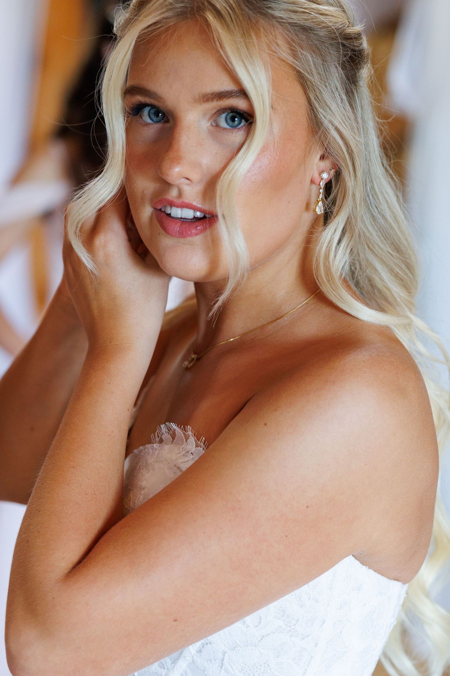 Blonde bride with blue eyes adjusting earring, wearing white lace dress and delicate gold jewelry.