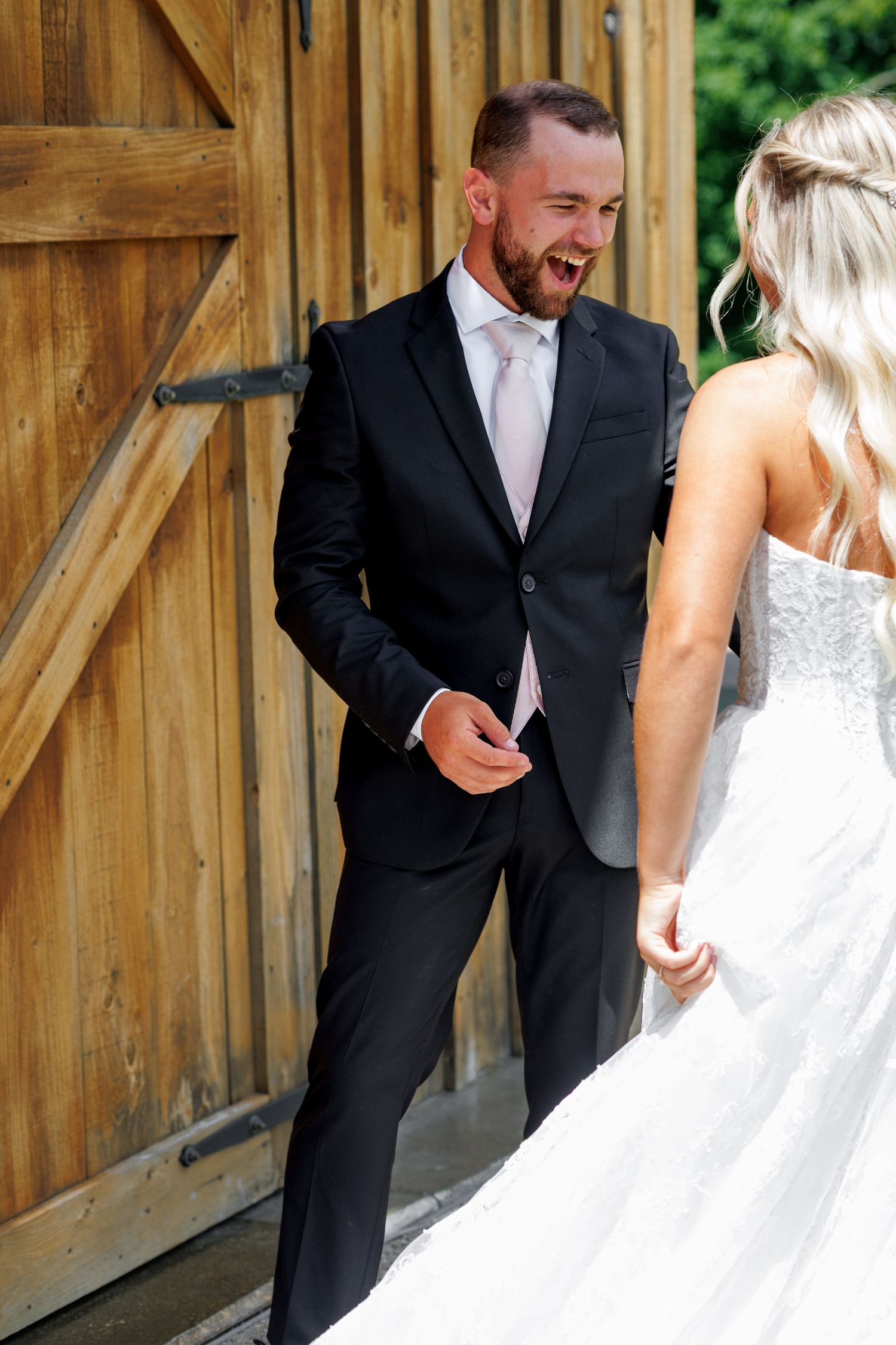 Groom in black suit laughing joyfully as bride in white lace gown approaches during wedding first look by barn doors.