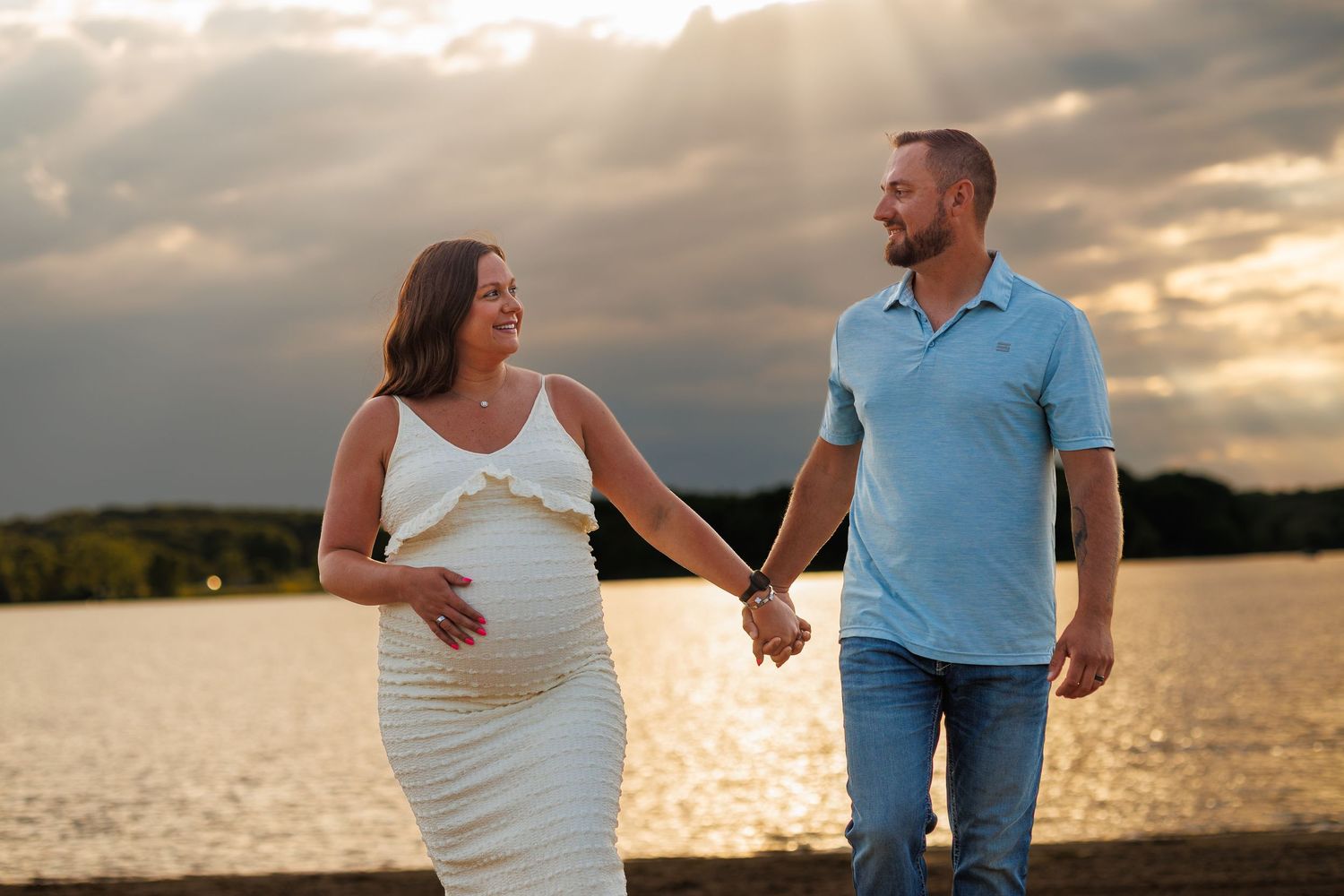 Pregnant couple holding hands by a lake at golden sunset, woman in white dress touching baby bump.