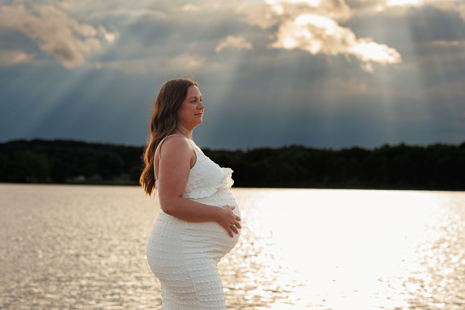 Pregnant woman in white dress standing by a lake with dramatic sunbeams breaking through clouds at golden hour.