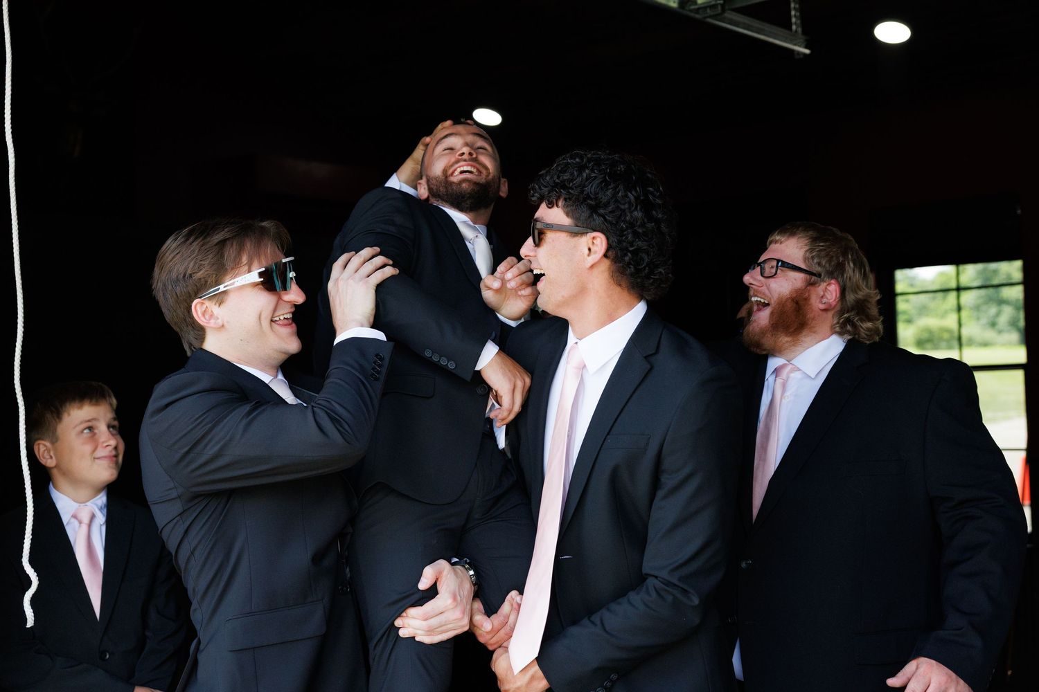 Groomsmen in black suits with pink ties laughing and joking together at a wedding celebration.