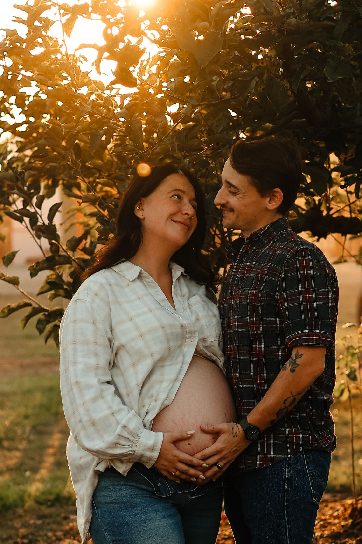 Happy pregnant couple gazing at each other tenderly under glowing sunset trees, both hands cradling baby bump.