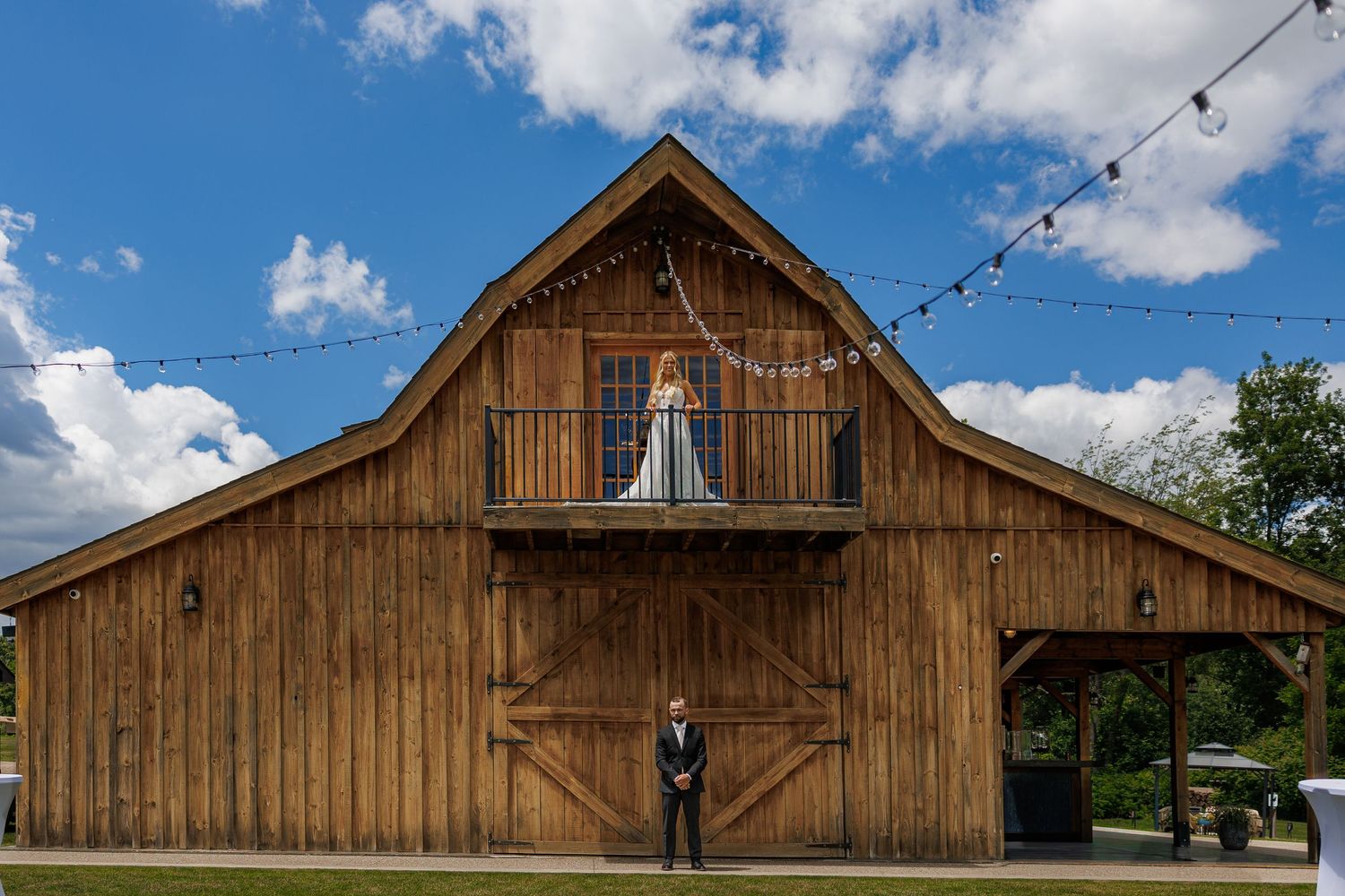 Bride on barn balcony with groom standing below at rustic wedding venue.