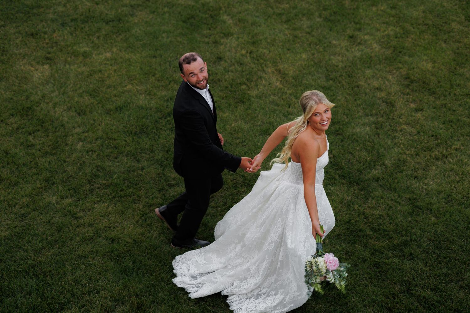 Aerial view of a bride and groom holding hands on green grass, bride in lace gown with bouquet, groom in black tuxedo.