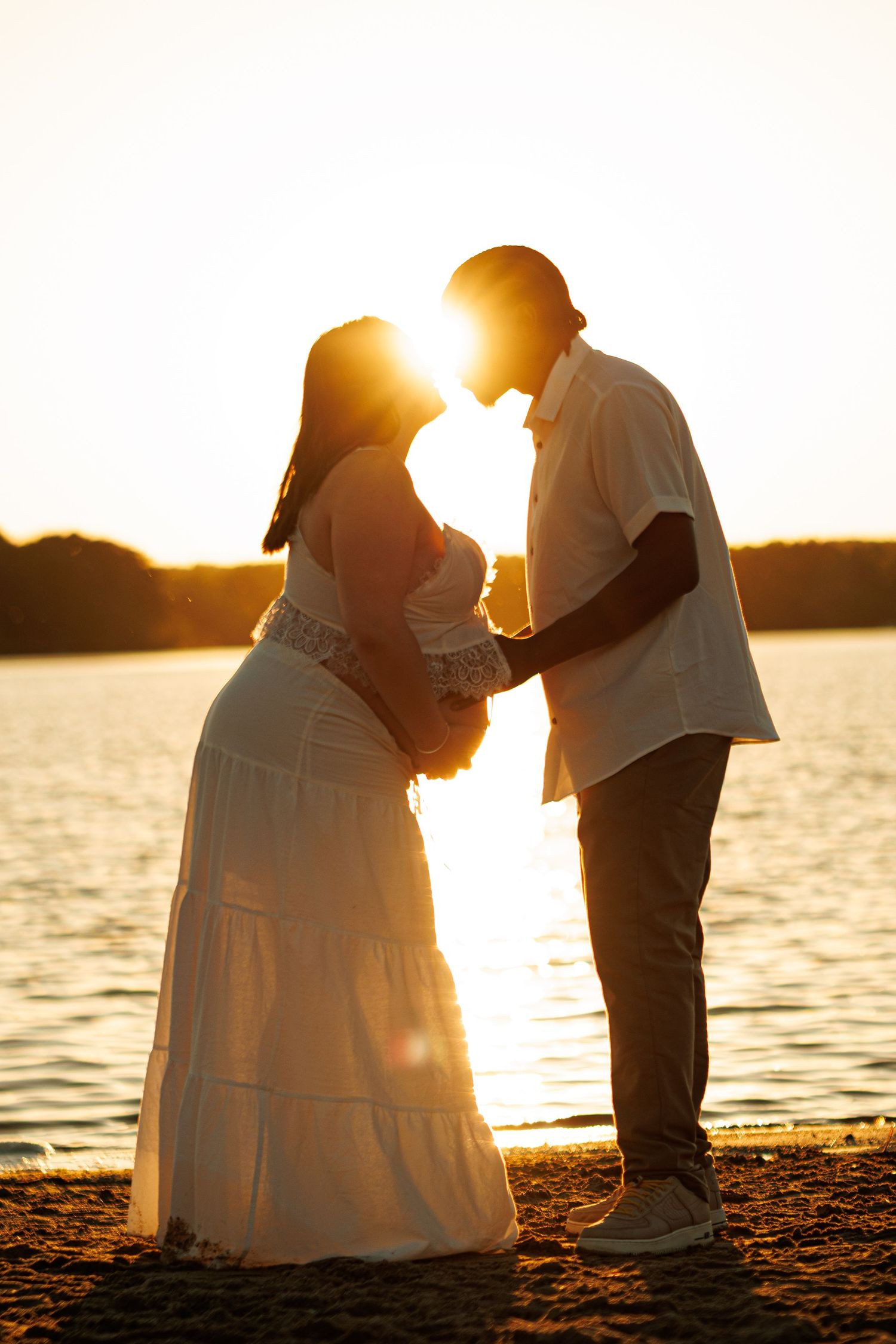 Pregnant couple silhouetted against golden sunset by the water, woman in white dress, man in casual attire, holding hands.