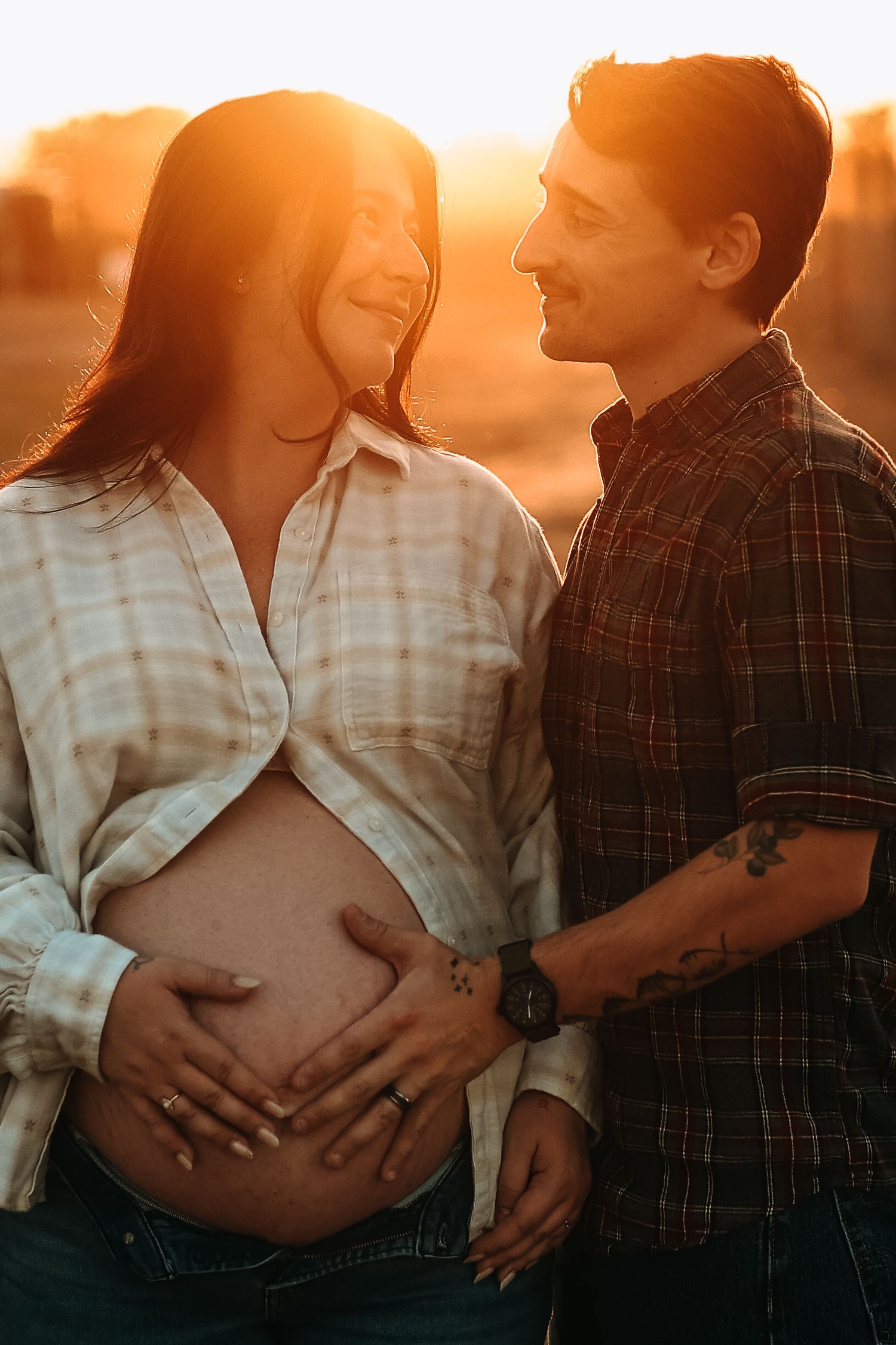 Pregnant couple embracing at sunset, woman in open plaid shirt showing baby bump with partners tattooed hand.
