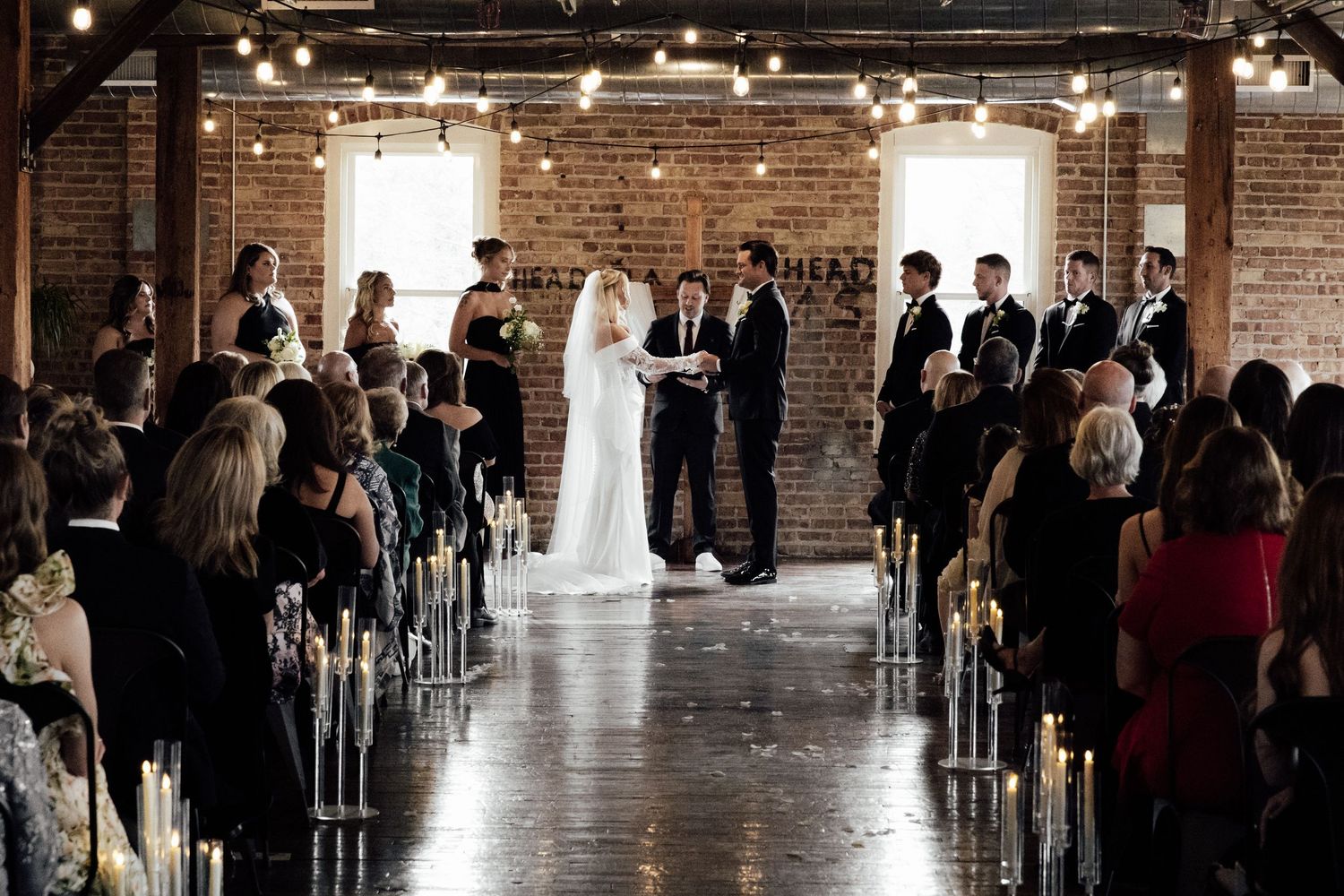 Wedding ceremony in rustic brick loft with string lights, bridal party at altar with seated guests.