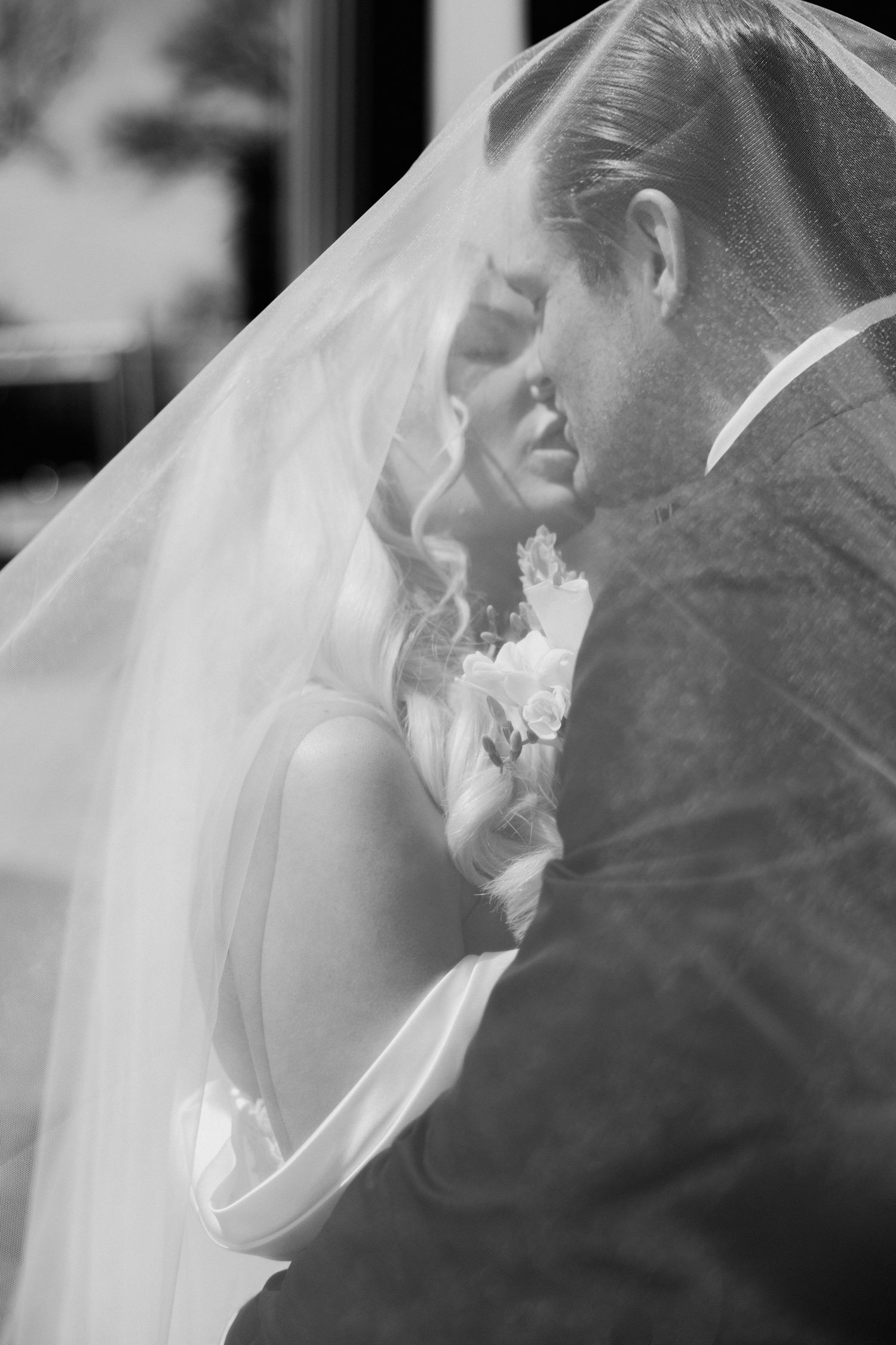 Black and white romantic photo of bride and groom beneath a sheer veil in soft light.