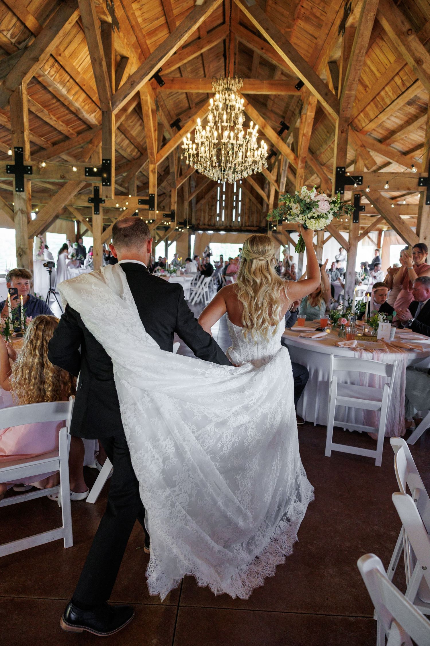 Bride and groom enter rustic barn wedding venue with chandeliers, guests seated at decorated tables.