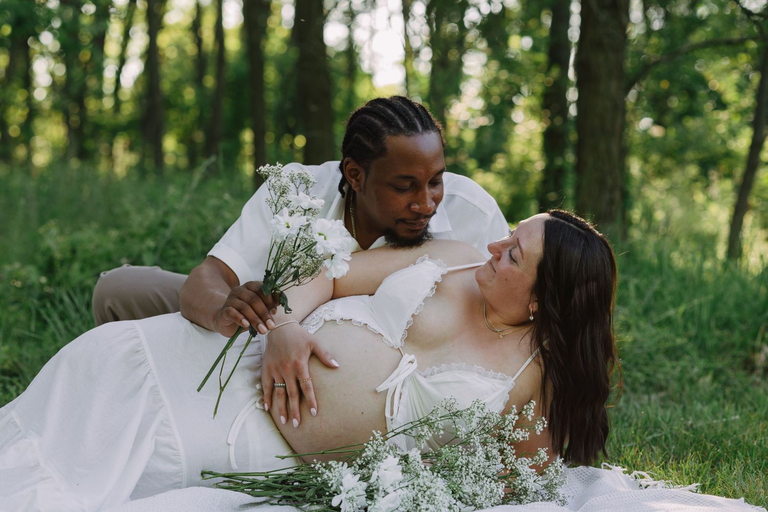 Couple shares tender maternity photo outdoors, man holding wildflowers beside pregnant woman in white dress among green trees.