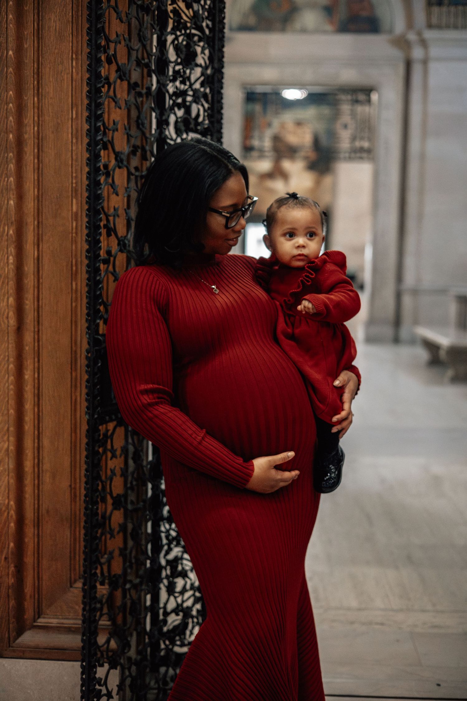 Pregnant mother in red ribbed dress holding baby girl in matching red outfit in ornate hallway.