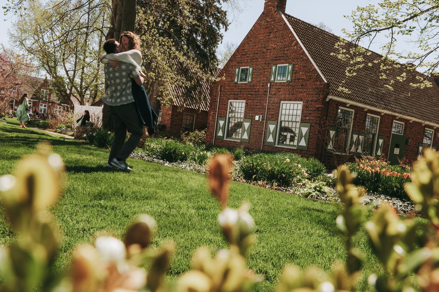 Couple embracing on a lush green lawn in front of a charming red brick house surrounded by spring flowers.