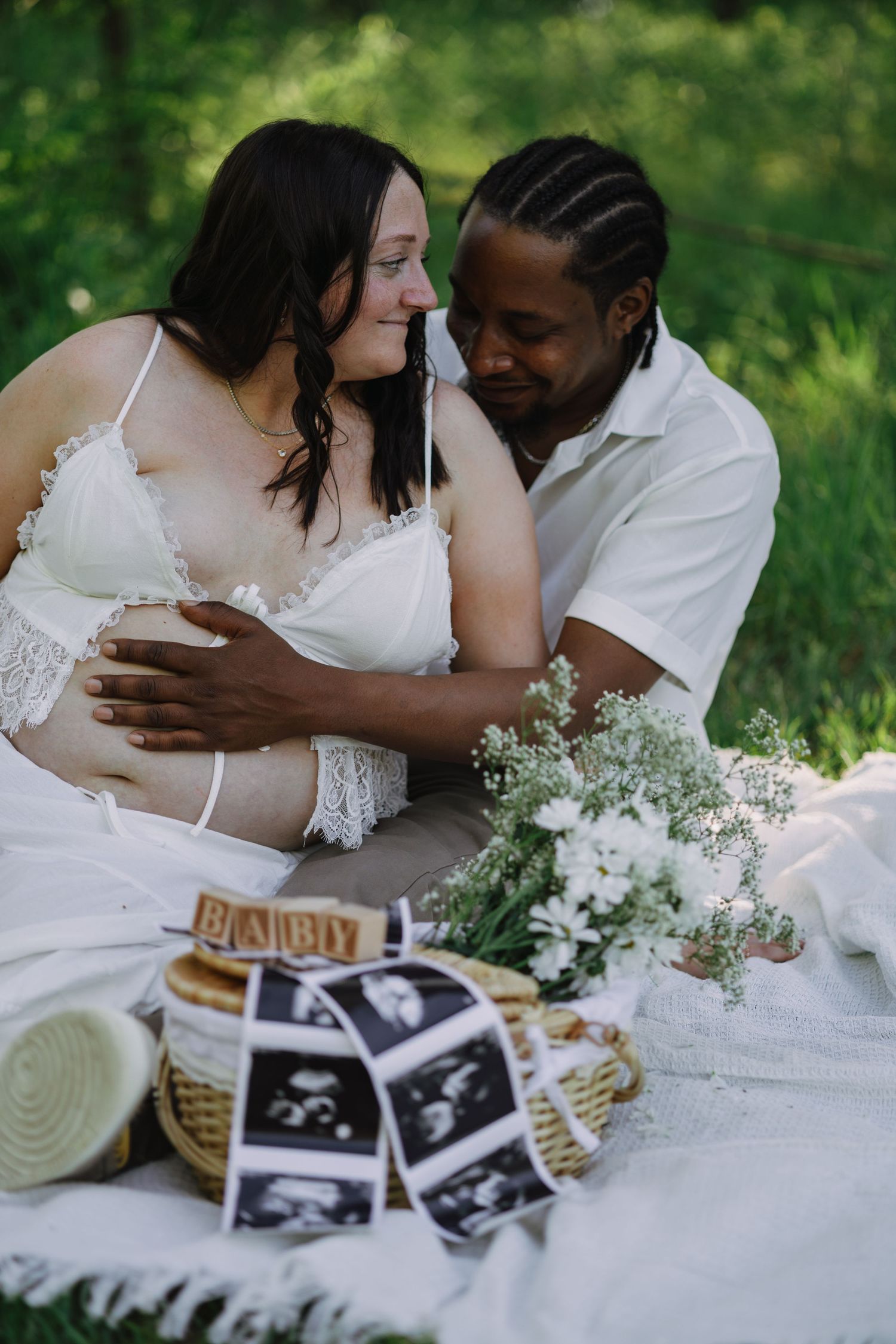 Expecting couple sitting on white blanket outdoors, with ultrasound photos and baby spelled blocks in foreground.