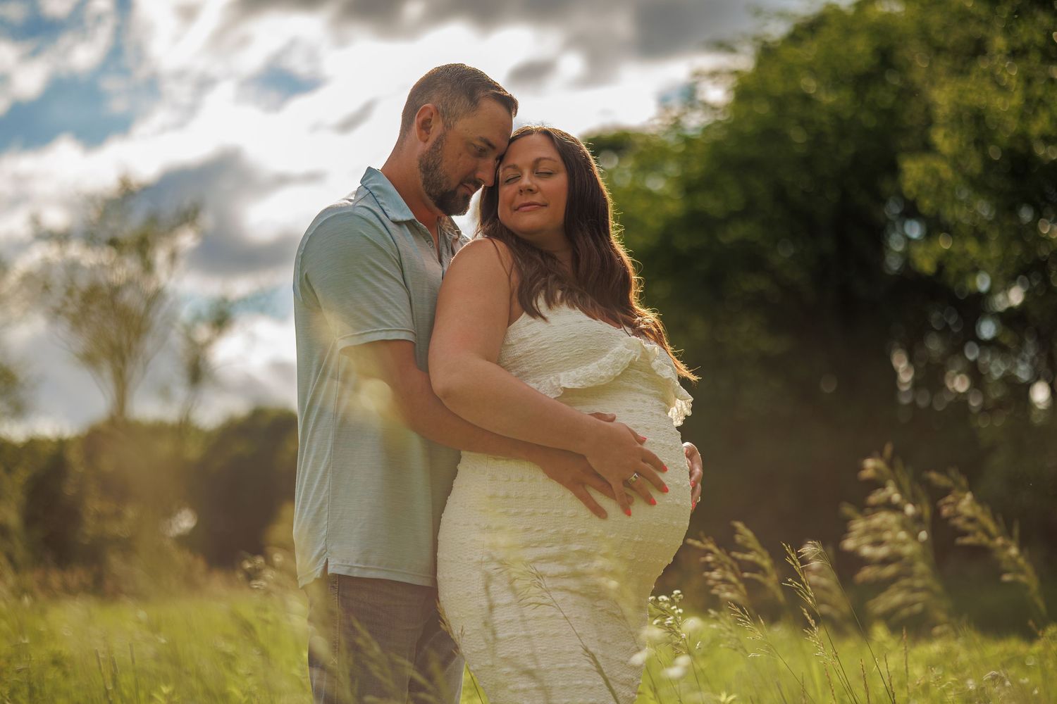 Couple embracing outdoors in sunny field, pregnant woman in white lace dress, man in blue shirt, maternity photo.