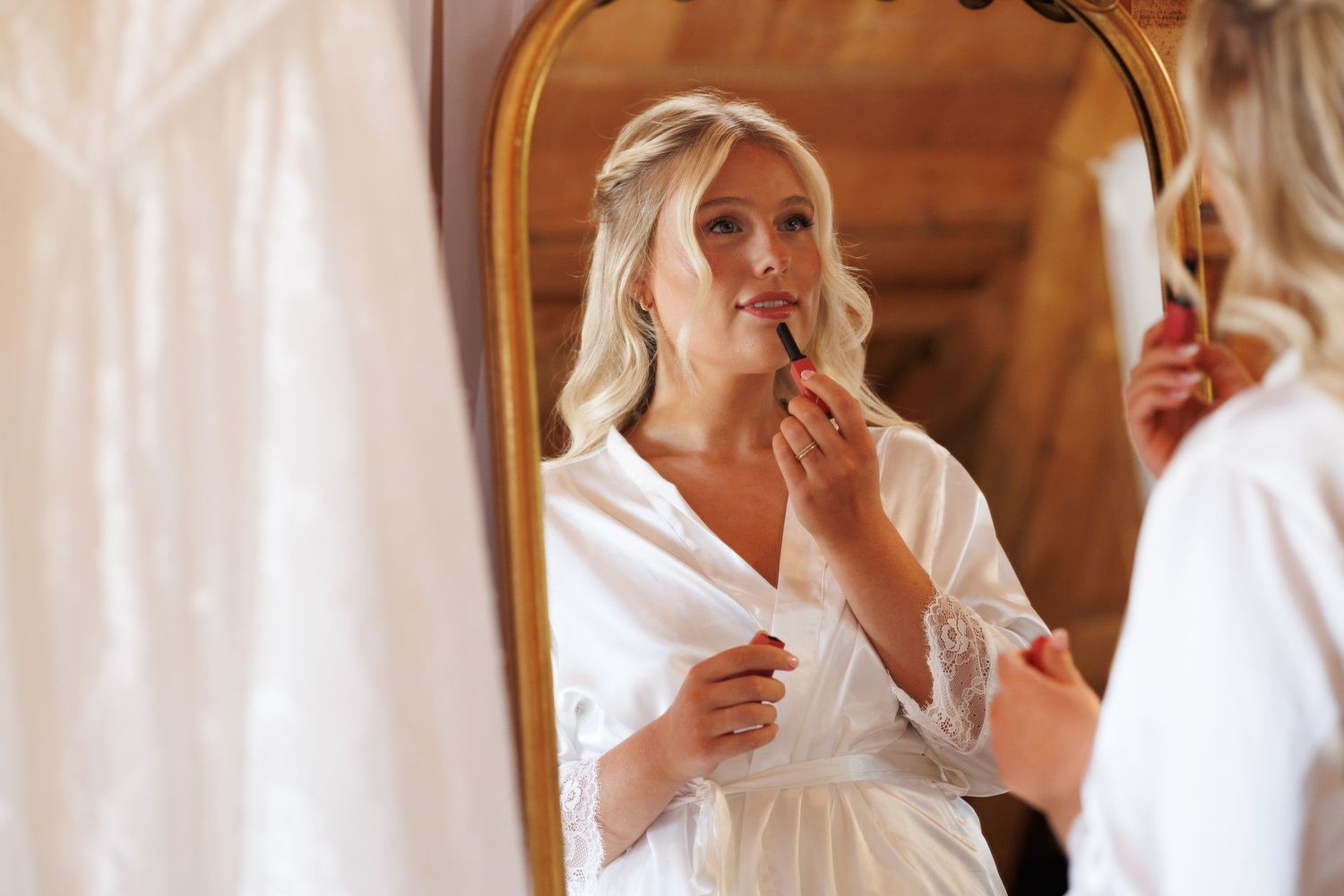 Bride in white lace robe applying lipstick while looking in ornate gold mirror during wedding preparations.