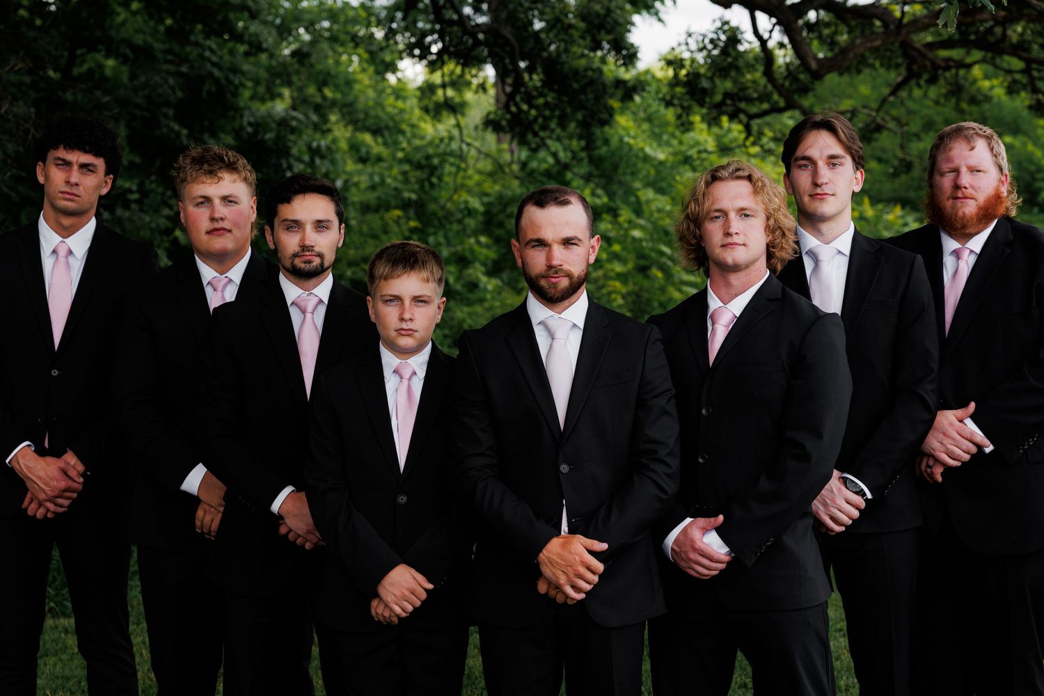 A group of seven groomsmen in matching black suits with pink ties pose outdoors for a formal wedding photo.
