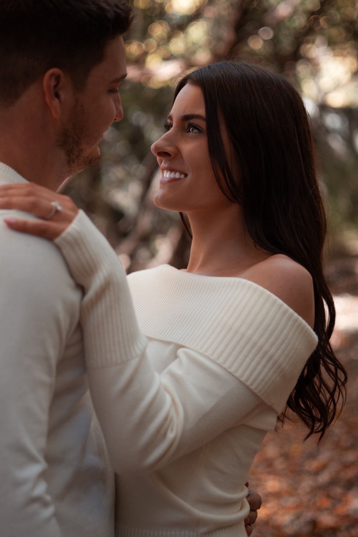 Happy woman in white off-shoulder sweater smiling at partner during fall engagement photo session in wooded setting.