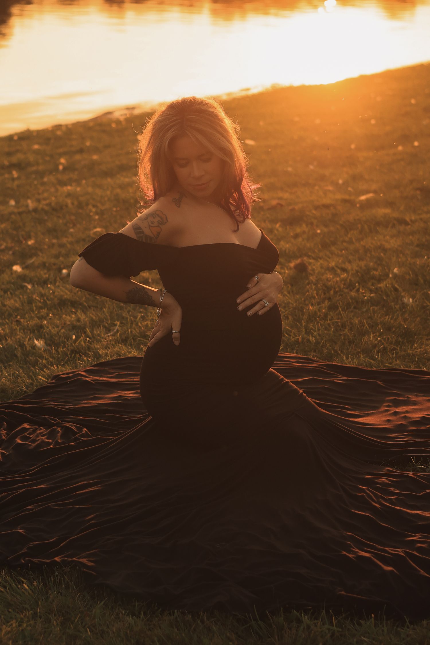 Pregnant woman in elegant black gown cradling belly by lakeside during golden hour sunset.
