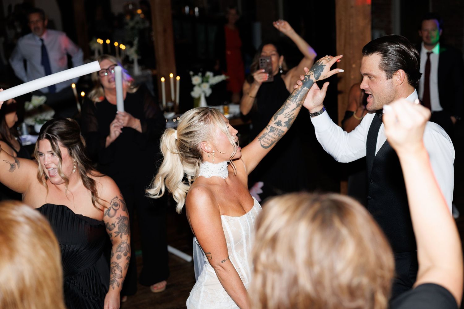 Tattooed bride in white gown dances energetically with groom and guests on crowded wedding reception floor.