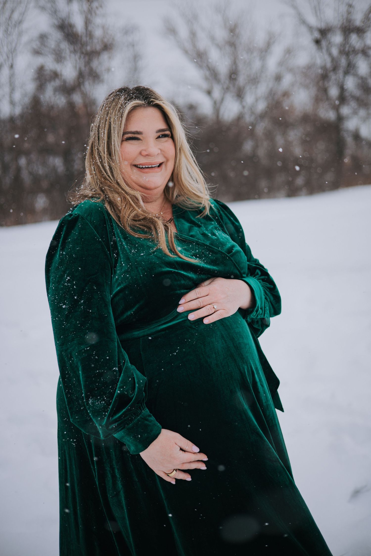 Glowing pregnant woman in green velvet wrap dress laughing while holding baby bump during snowy maternity session.