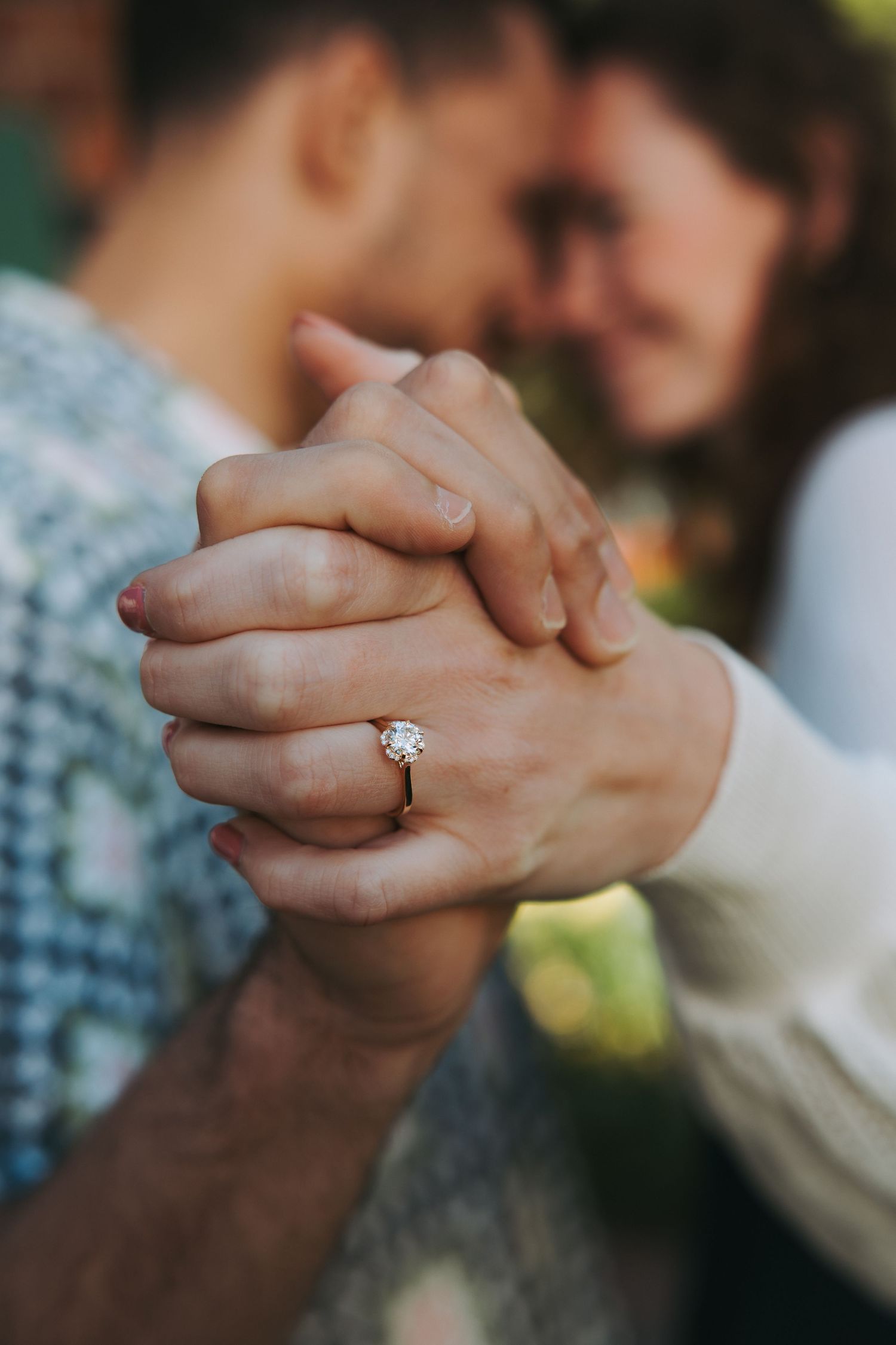 Engaged couple holding hands closely showing diamond solitaire ring while leaning foreheads together.