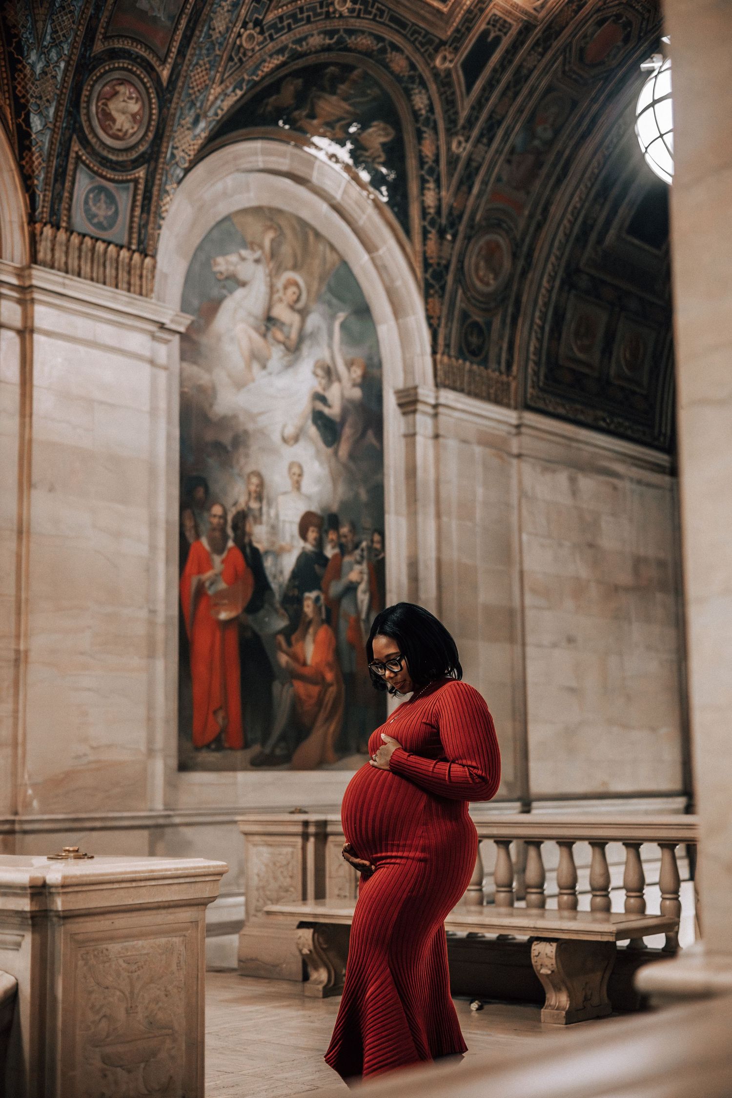 Pregnant woman in red ribbed dress cradling baby bump inside ornate church with Renaissance paintings.