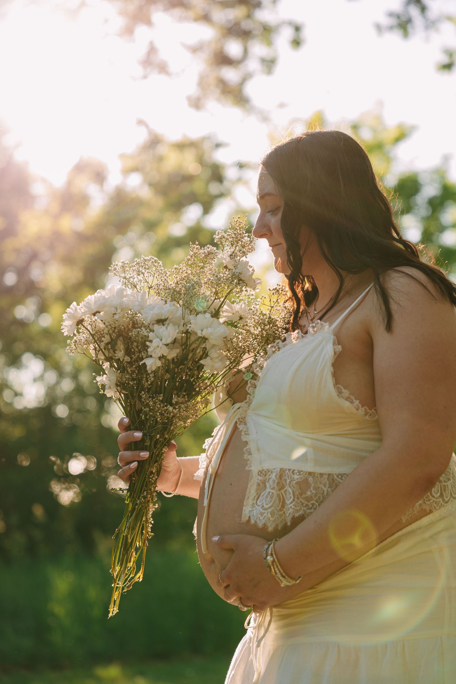 Pregnant woman in yellow dress holding white wildflowers bouquet, glowing in golden sunlight outdoors.