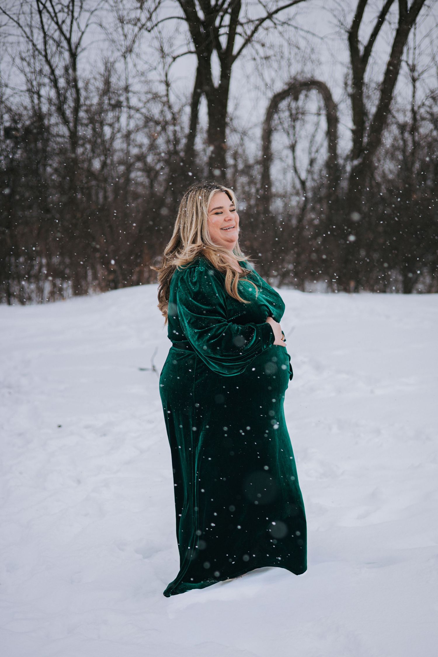 Pregnant woman in elegant emerald velvet gown posing gracefully in snowy winter landscape during maternity photoshoot.
