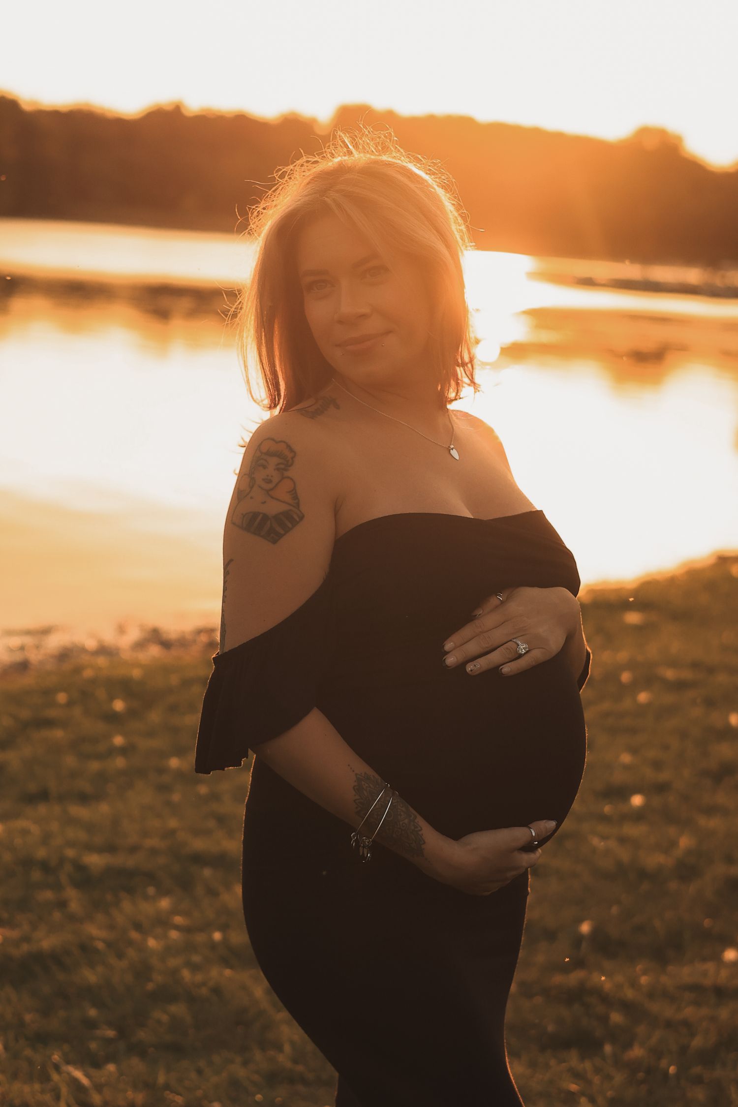 Tattooed pregnant woman in black off-shoulder dress holding belly at golden hour lakeside.
