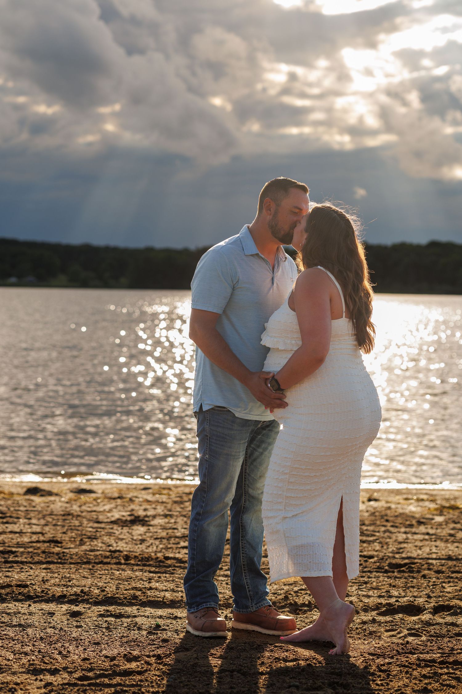 Couple kissing on a sandy beach at sunset with dramatic cloudy sky and shimmering lake in the background.