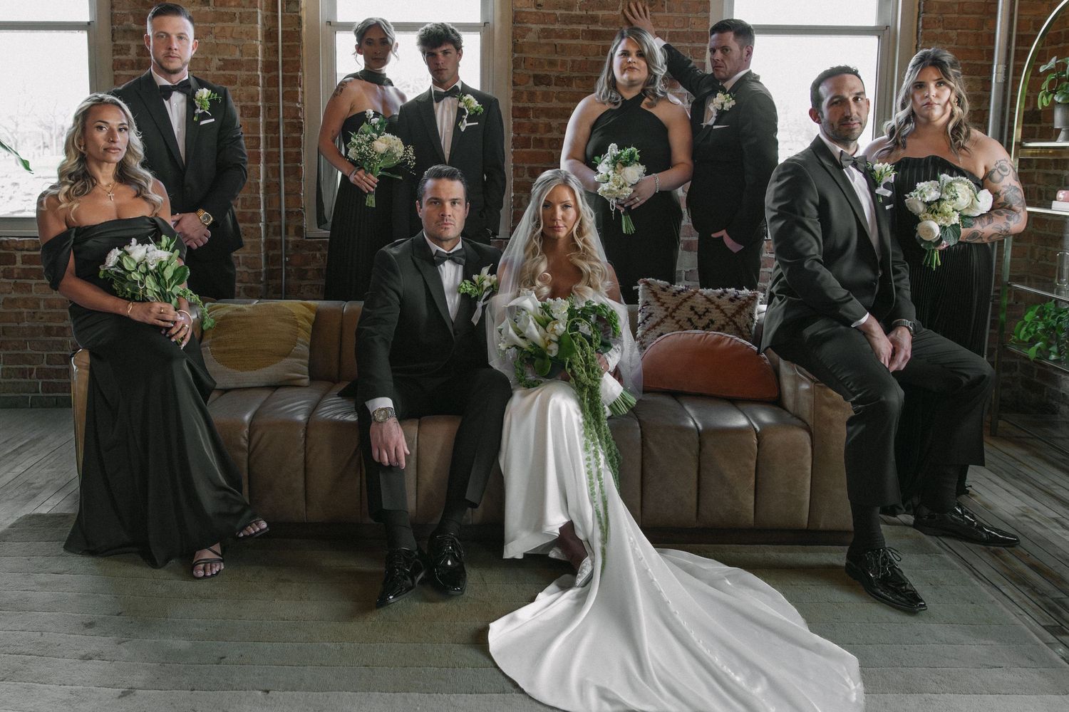 Wedding party poses together in black formal attire with white floral bouquets in industrial venue.