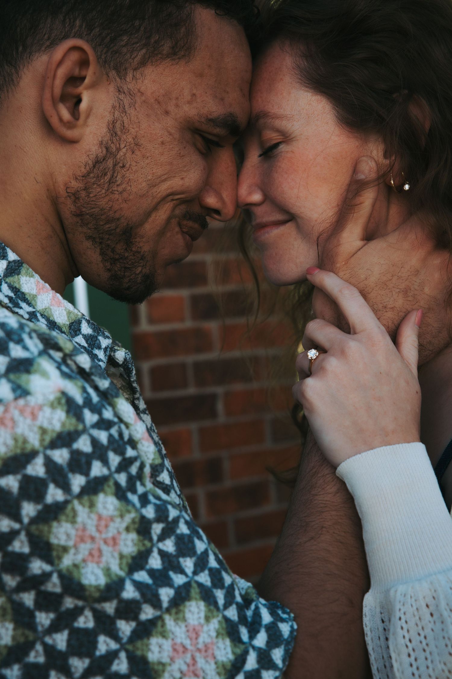 Intimate close-up of a couple touching foreheads with woman showing engagement ring against a brick wall backdrop.
