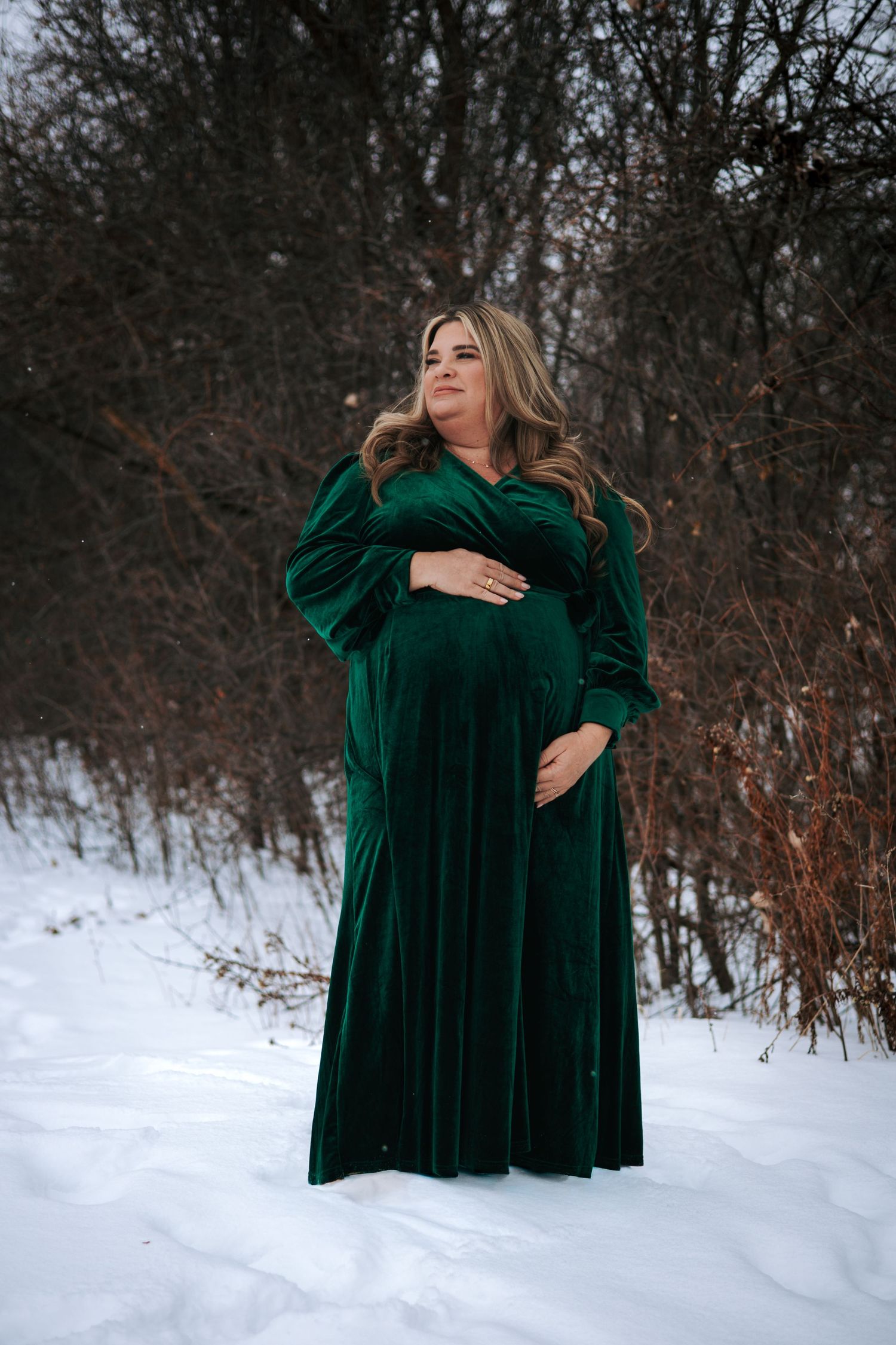 Pregnant woman in elegant green velvet maternity gown standing in snowy winter landscape.
