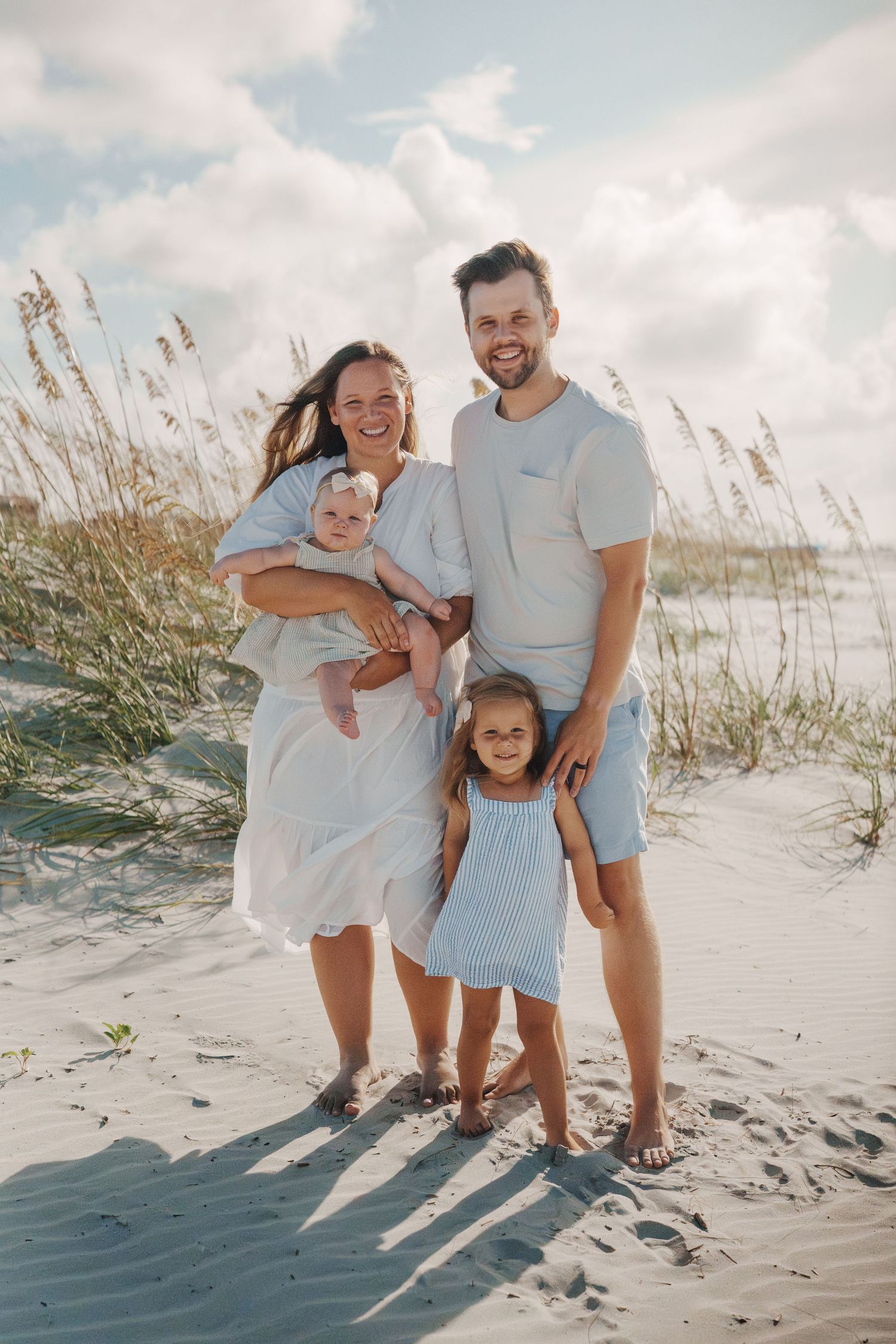Happy family of four poses on sandy beach with sea oats and cloudy sky.
