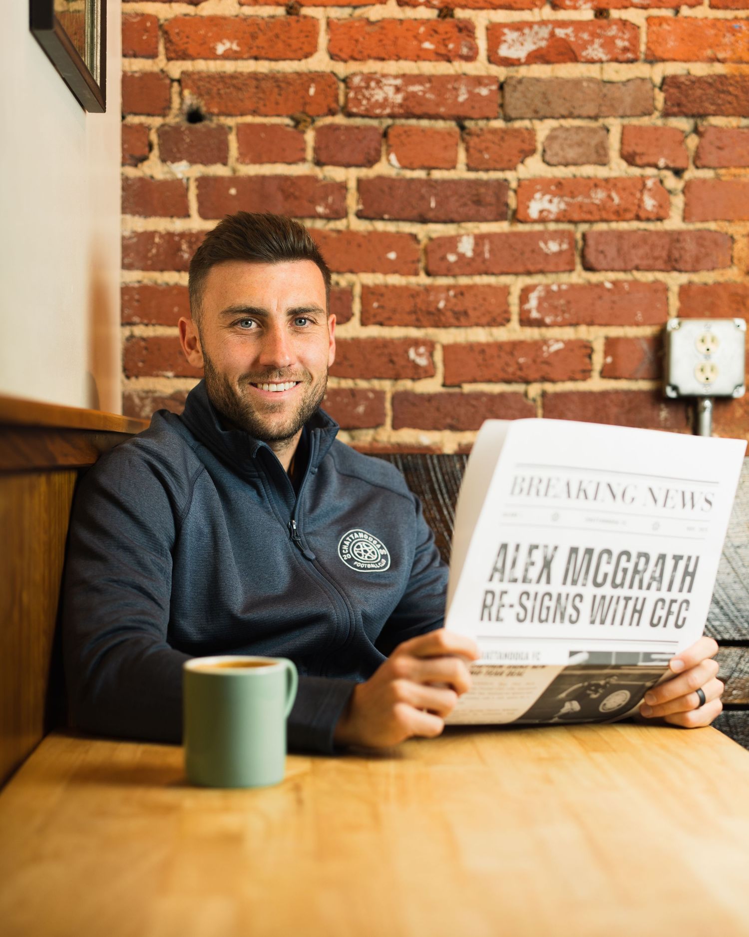 Smiling man holds Alex McGrath CFC re-signing newspaper in rustic brick interior cafe. Photographed for Chattanooga FC player signing.
