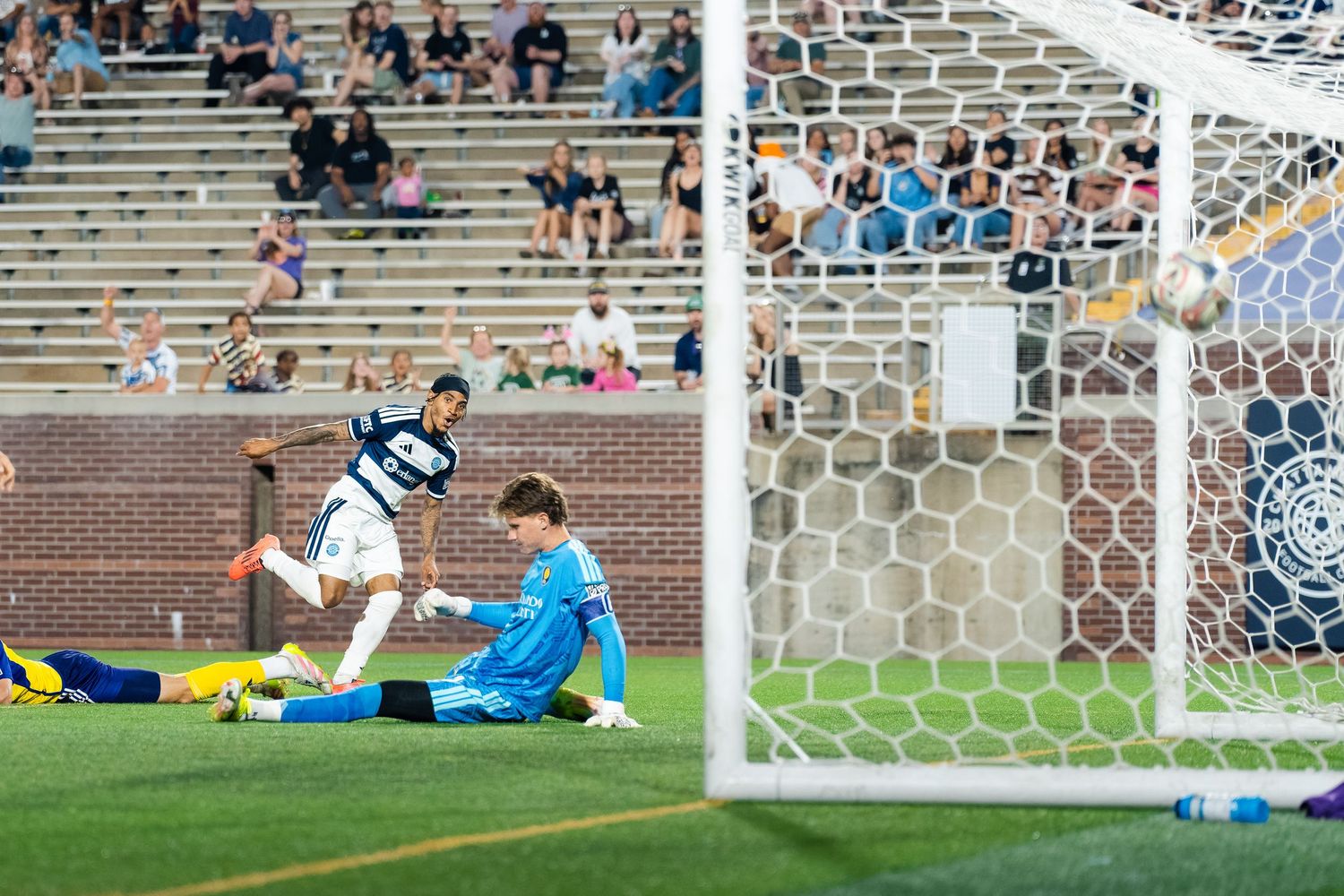 Chattanooga FC Soccer player scores goal past diving goalkeeper during competitive MLS Next Pro match in stadium setting.