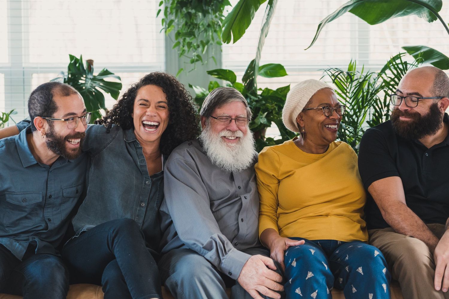 A group of five adults laugh together joyfully on a couch with green plants.