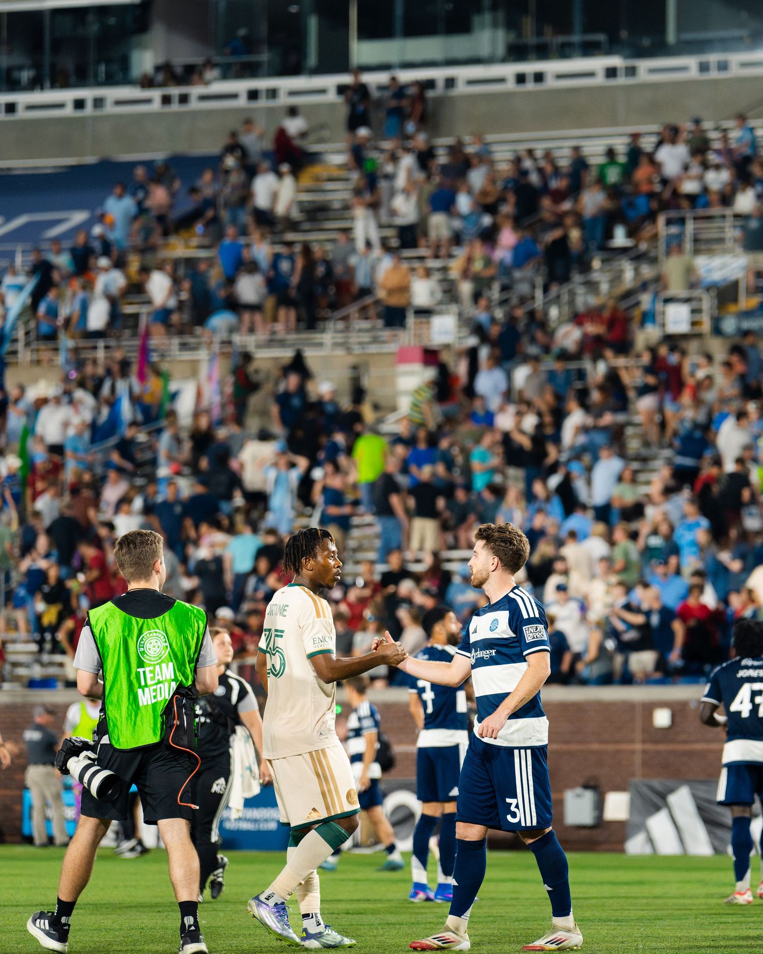 Soccer players shake hands after match as large crowd fills stadium stands behind them.