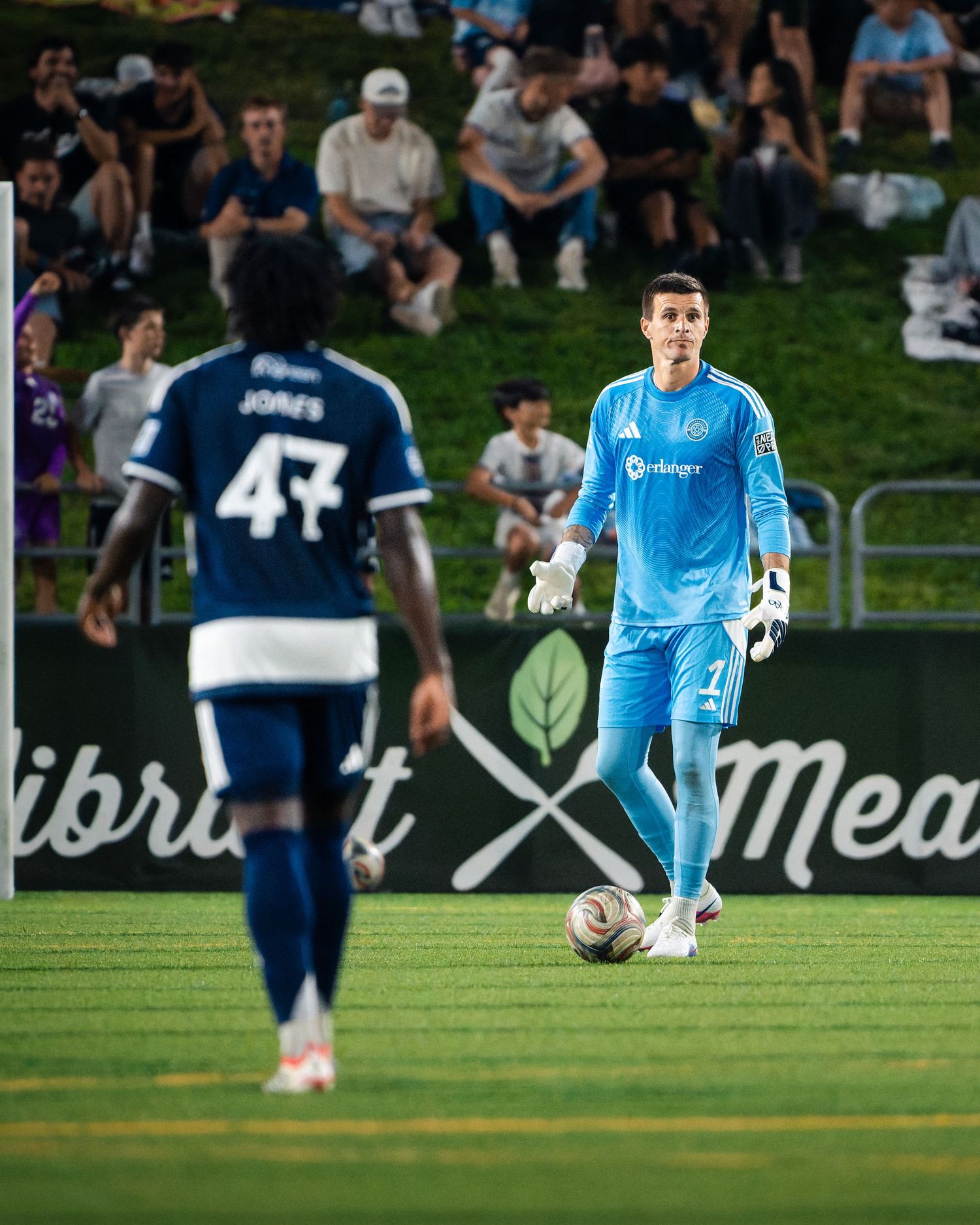 Chattanooga FC Soccer goalkeeper in light blue kit controls ball while teammate wearing number 47 stands nearby.
