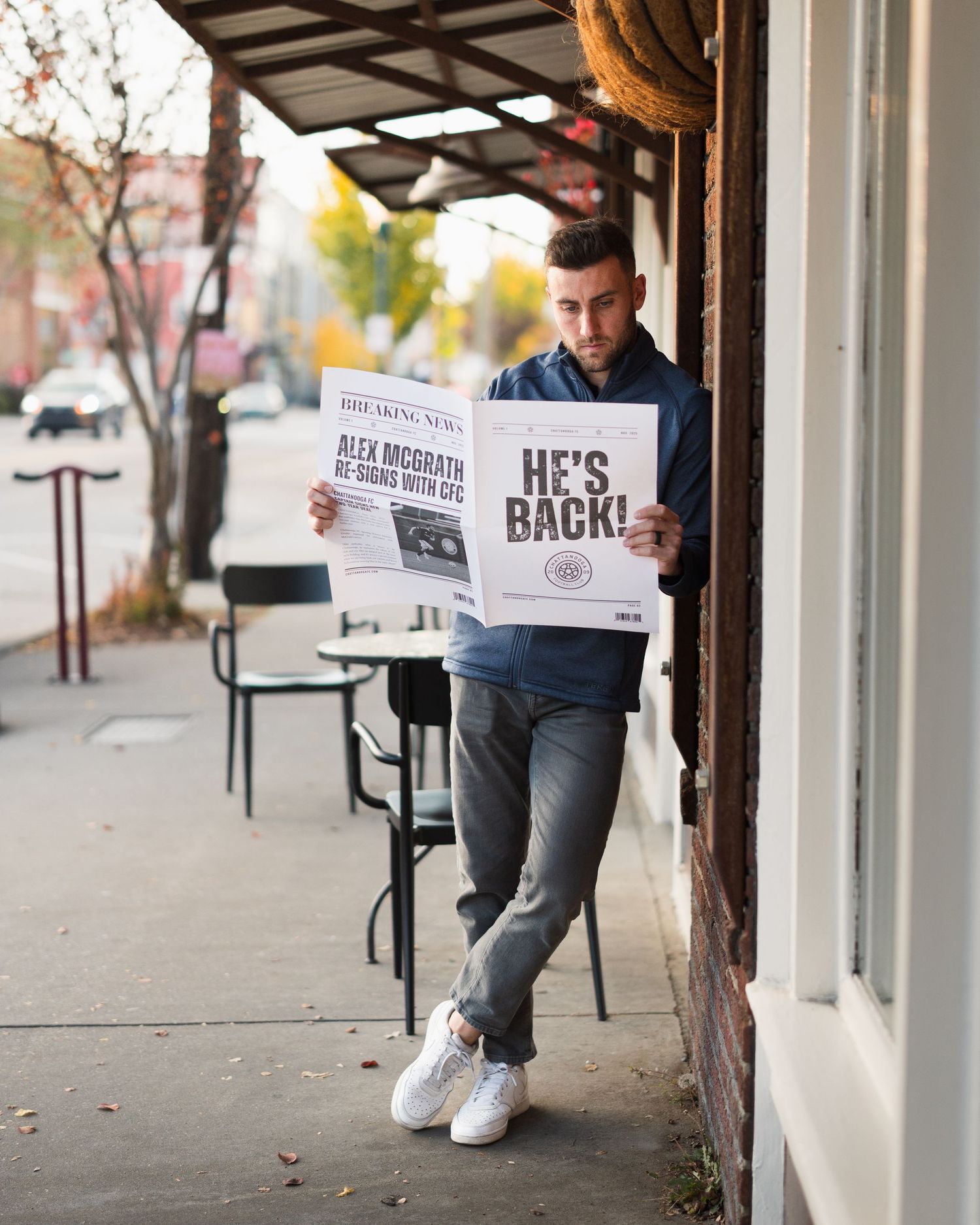 Man in hoodie reads HE'S BACK newspaper leaning against building on autumn sidewalk. Photographed for Chattanooga FC player signing.